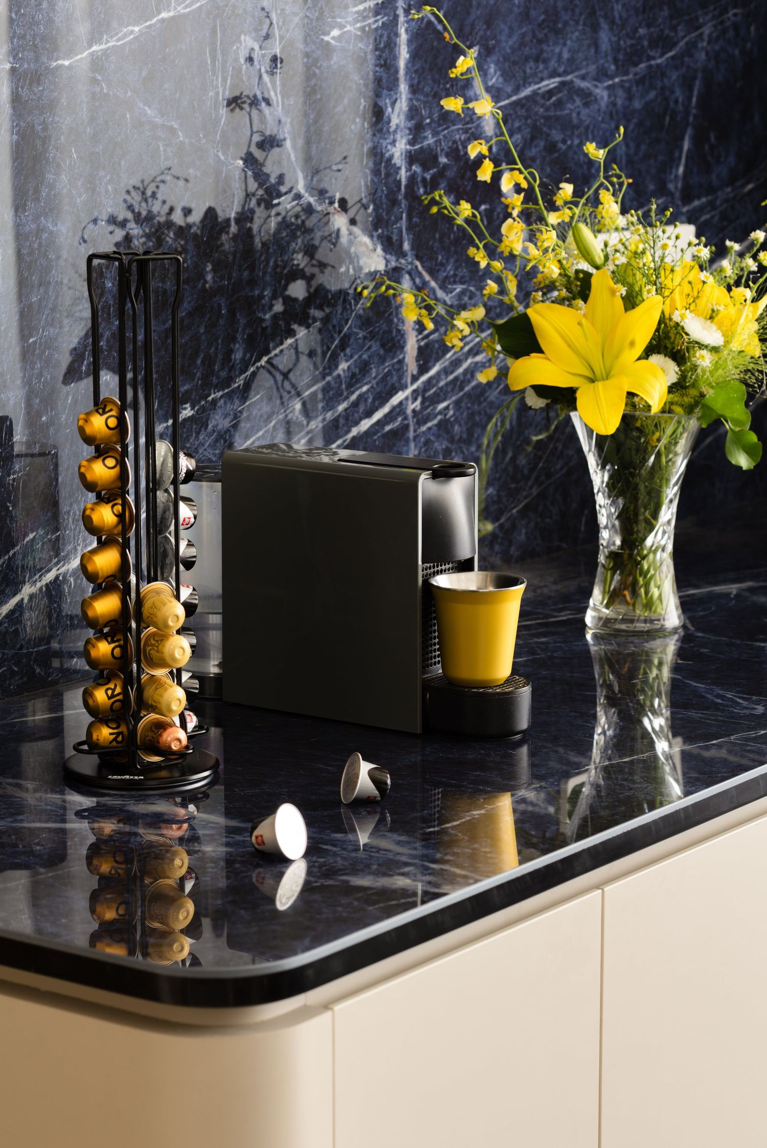 Modern black coffee station with yellow lilies in vase, coffee pod holder, and espresso machine on marble countertop.