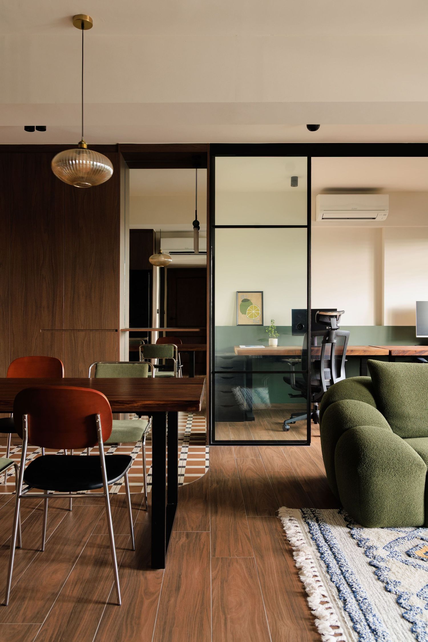 Modern apartment interior with glass partition, green sofa, orange dining chairs and wood flooring under pendant lighting.