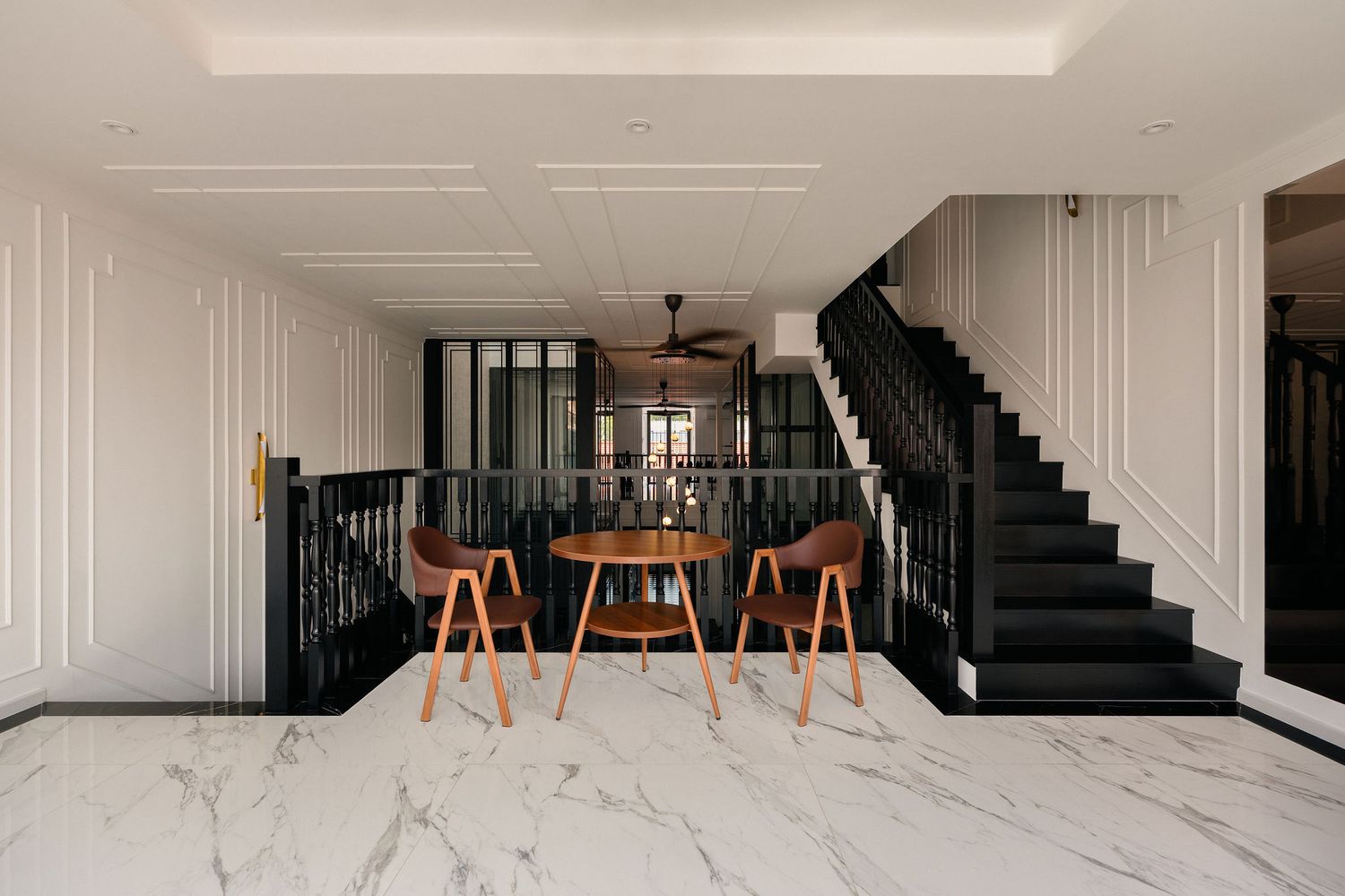 Elegant interior with white marble flooring, a black staircase with wainscoting walls, and a round wooden table with two mid-century chairs.