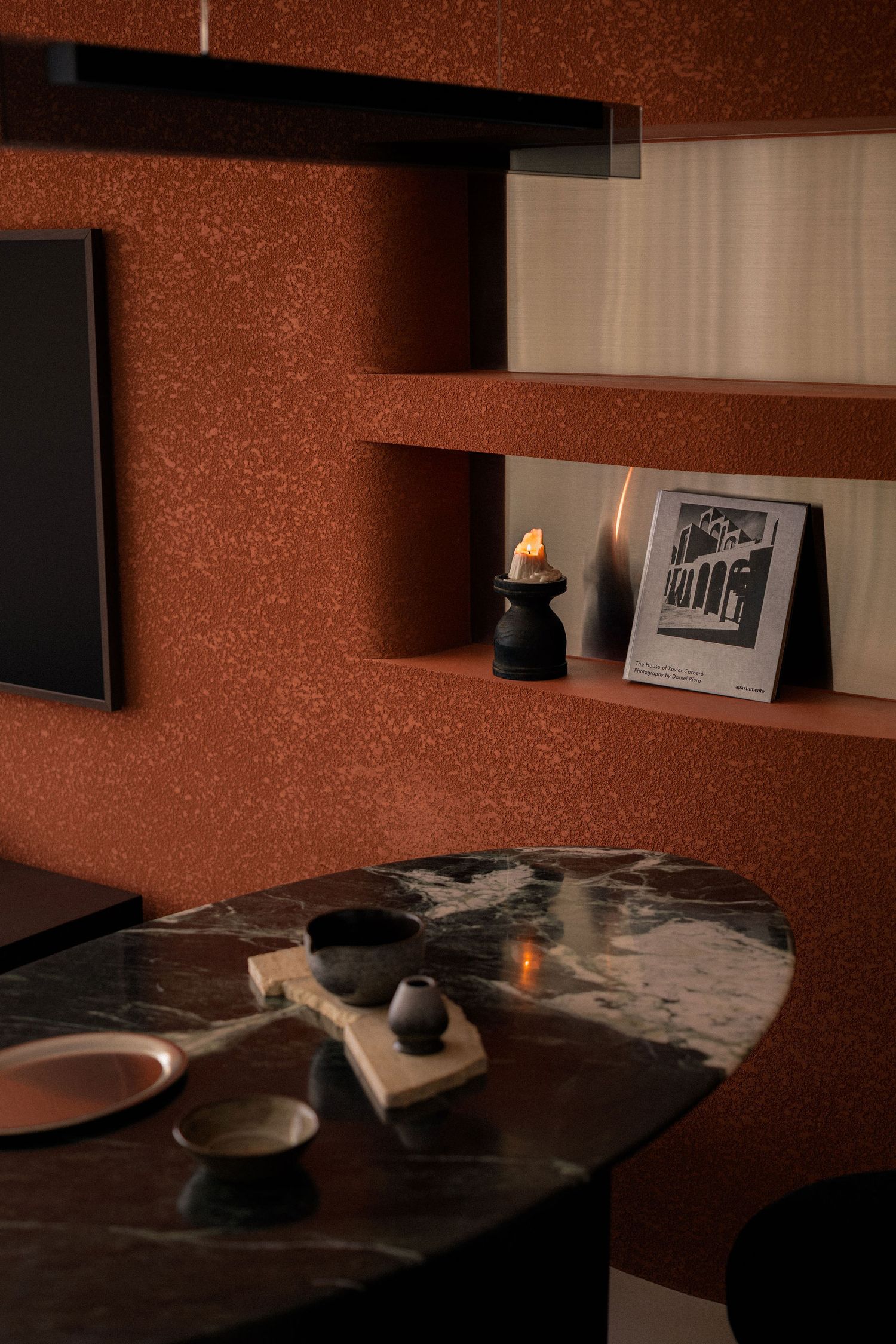 Terracotta-textured wall with built-in shelves, a framed print and candle above a black marble oval table with ceramic bowls and a tray.
