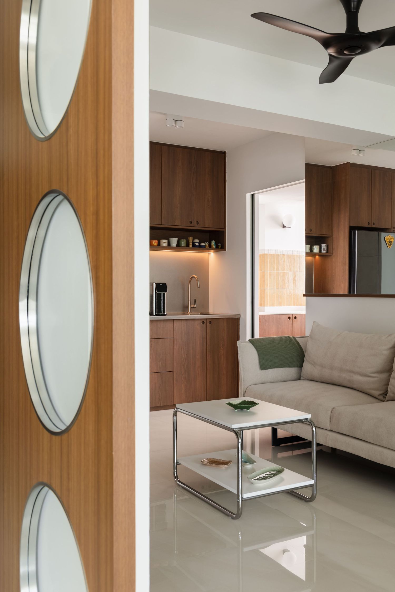 Modern apartment interior featuring wooden cabinetry, a grey sofa, glass coffee table, and ceiling fan with decorative oval door panels.