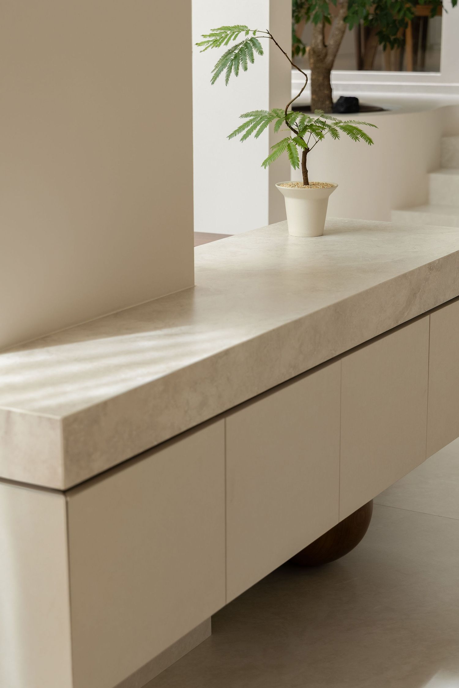 Minimalist cream marble sideboard with cabinet doors, topped with a small potted green plant near a sunlit window.
