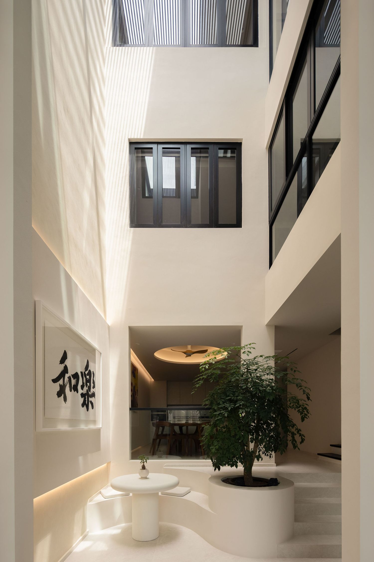 Modern atrium with Japanese calligraphy wall art, indoor tree, white furniture, and multi-story windows flooding space with natural light.