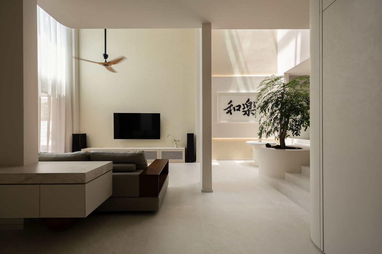 Minimalist living room with neutral tones, wall-mounted TV, ceiling fan, and an indoor bonsai tree near a sunlit alcove.