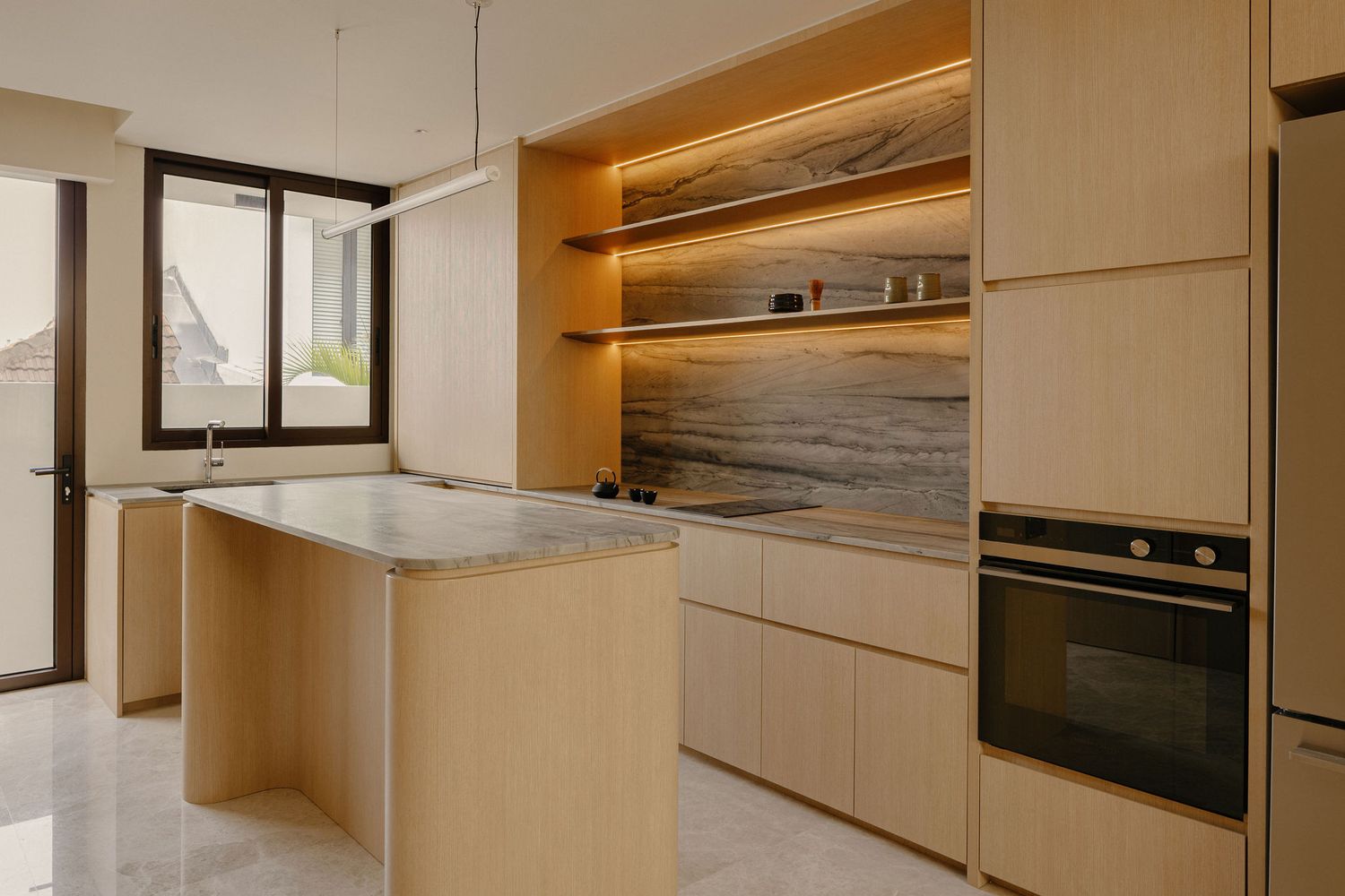 Modern kitchen with light wood cabinetry, marble island, built-in oven, and warm LED-lit open shelving.