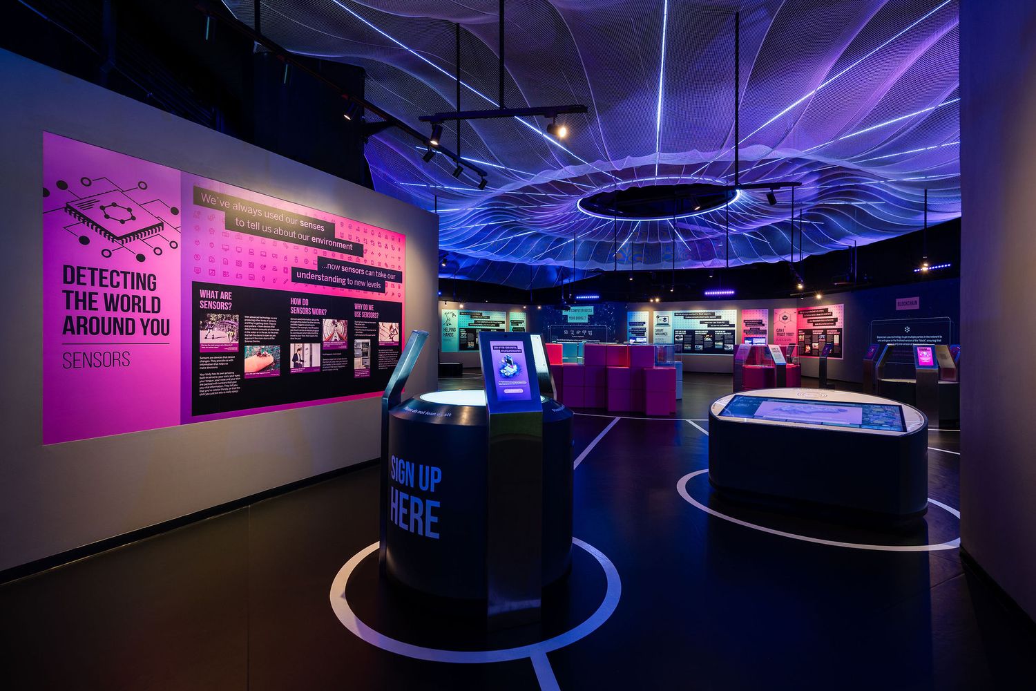 Interactive science museum exhibit with neon purple lighting, digital displays, and futuristic ceiling installation.