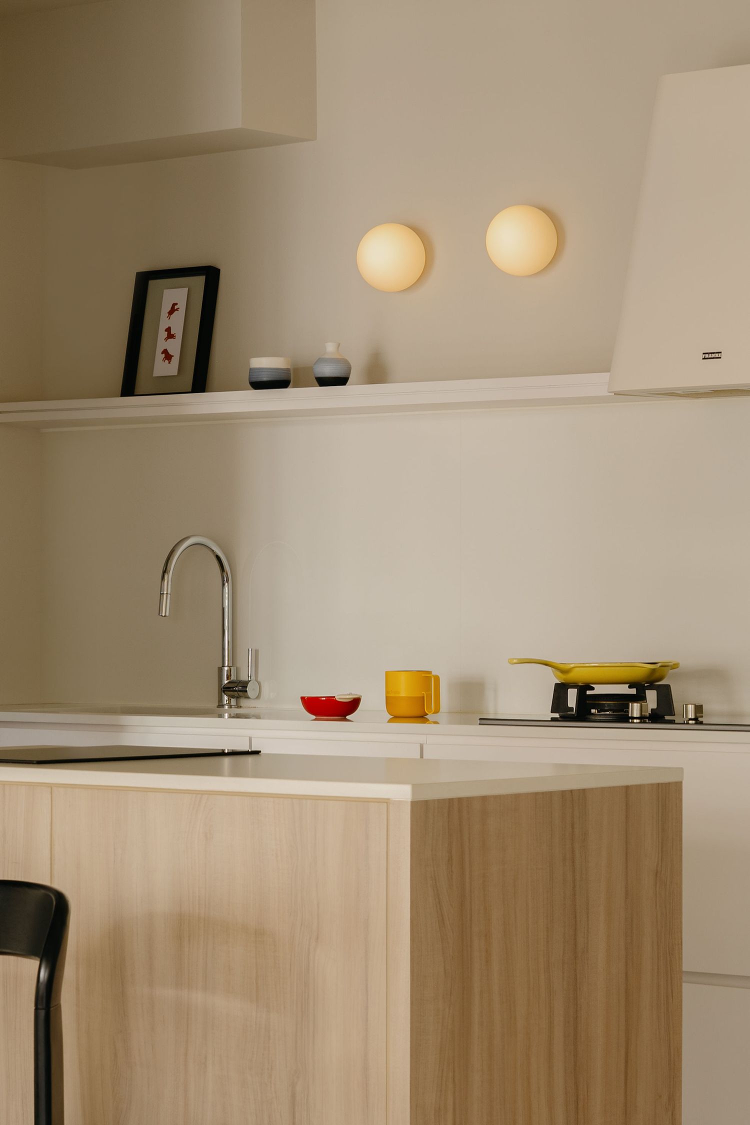Modern minimalist kitchen with wood island, chrome faucet, colorful mugs, globe wall lights, and floating shelves.