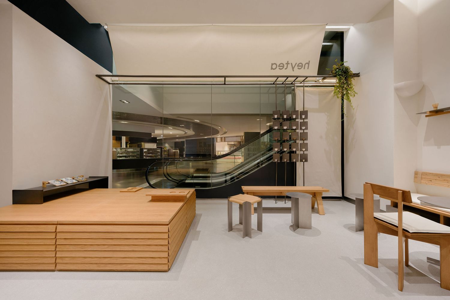 Minimalist retail interior with light wood platform, wooden stools and benches, glass storefront and escalator view beyond.