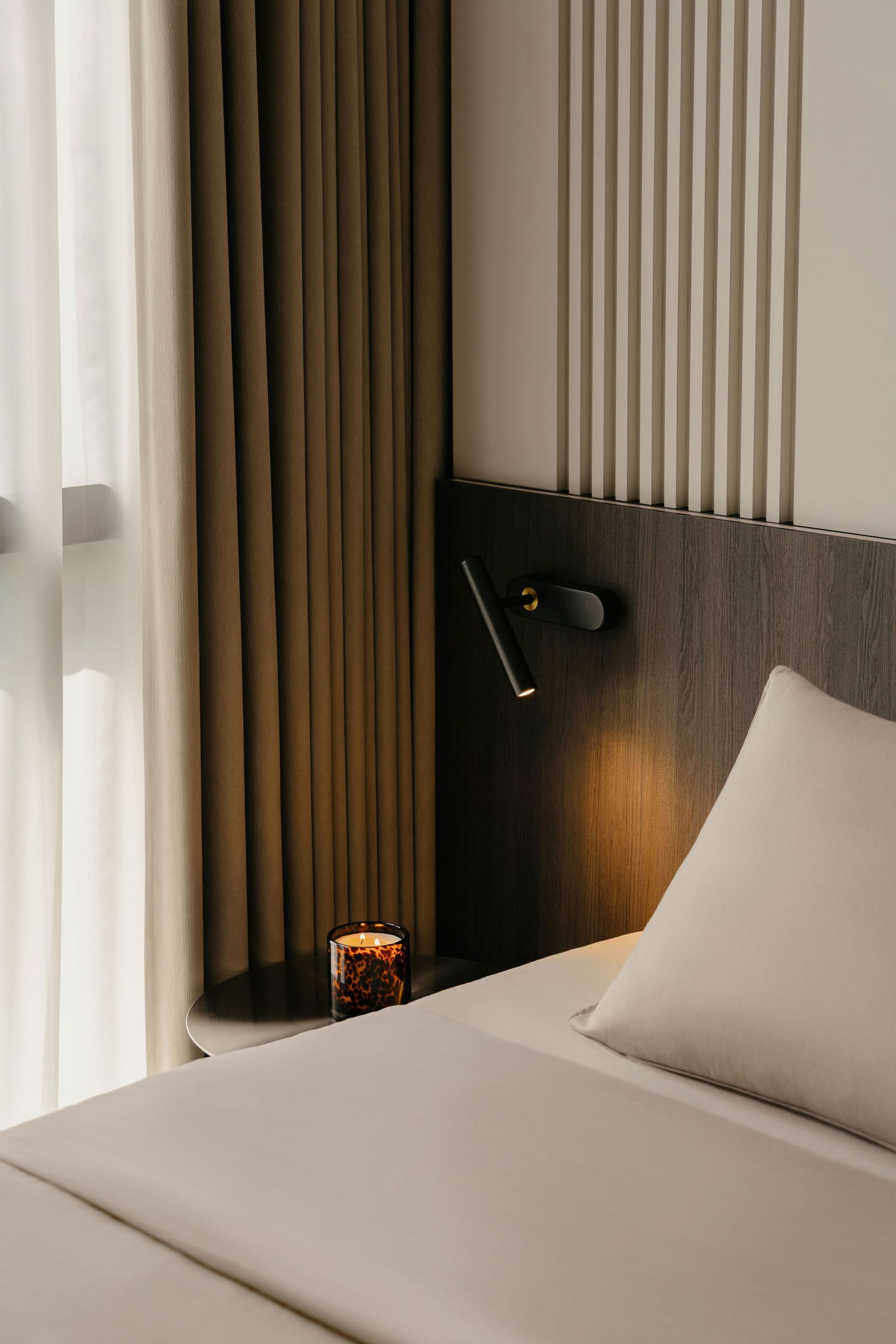 Cozy hotel bedroom corner with a wall-mounted reading light, lit candle on a round bedside table, beige linens and warm curtains.