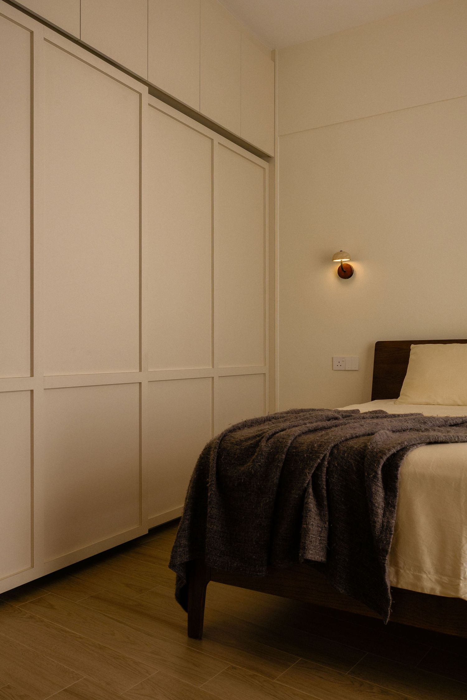 Minimalist bedroom with cream built-in wardrobes, a wooden bed draped in a gray knit blanket, and a warm wall sconce.