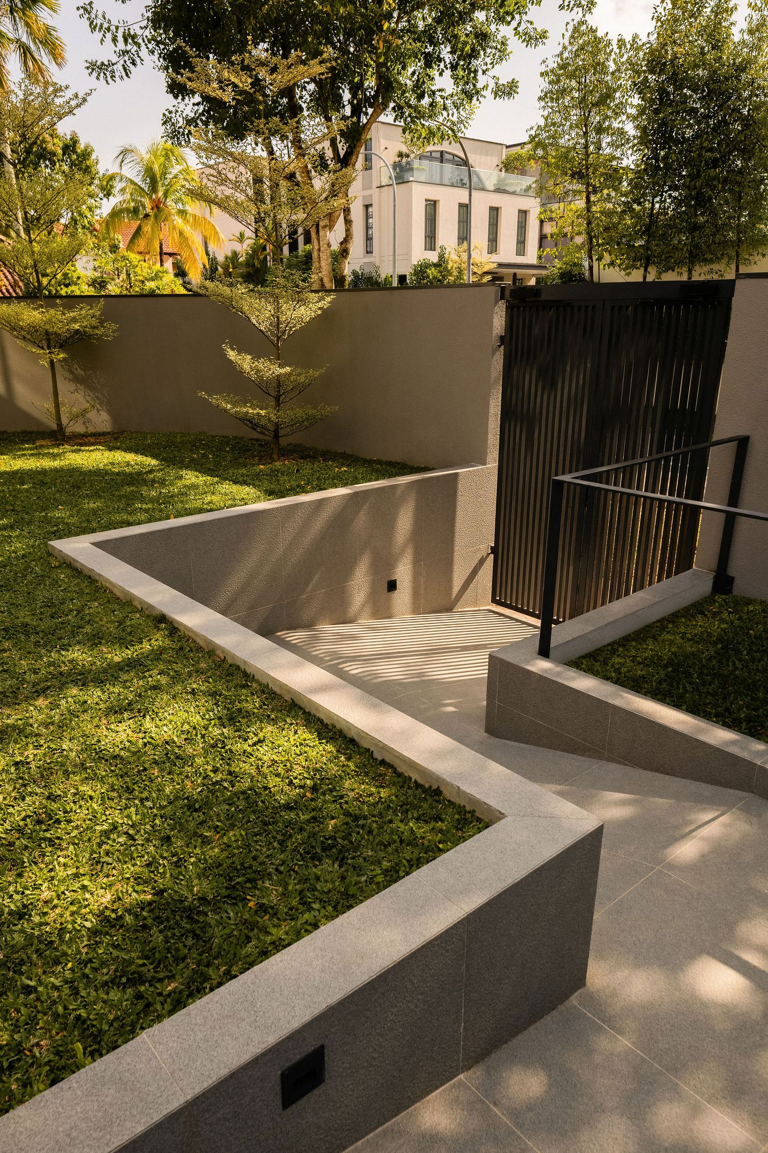 Modern sunken garden with a concrete ramp, black metal railing and slatted gate beside a lush green lawn and tropical trees.