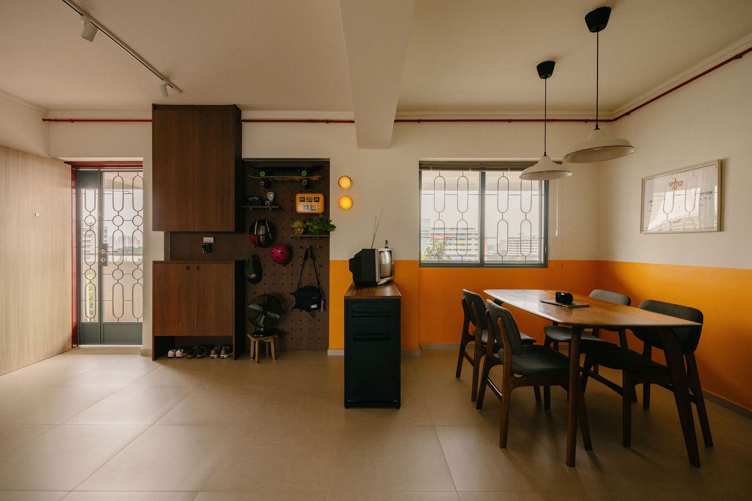 Bright open-plan dining area with a dark wood table, black chairs, orange accent walls, pendant lamps, and a pegboard display wall.