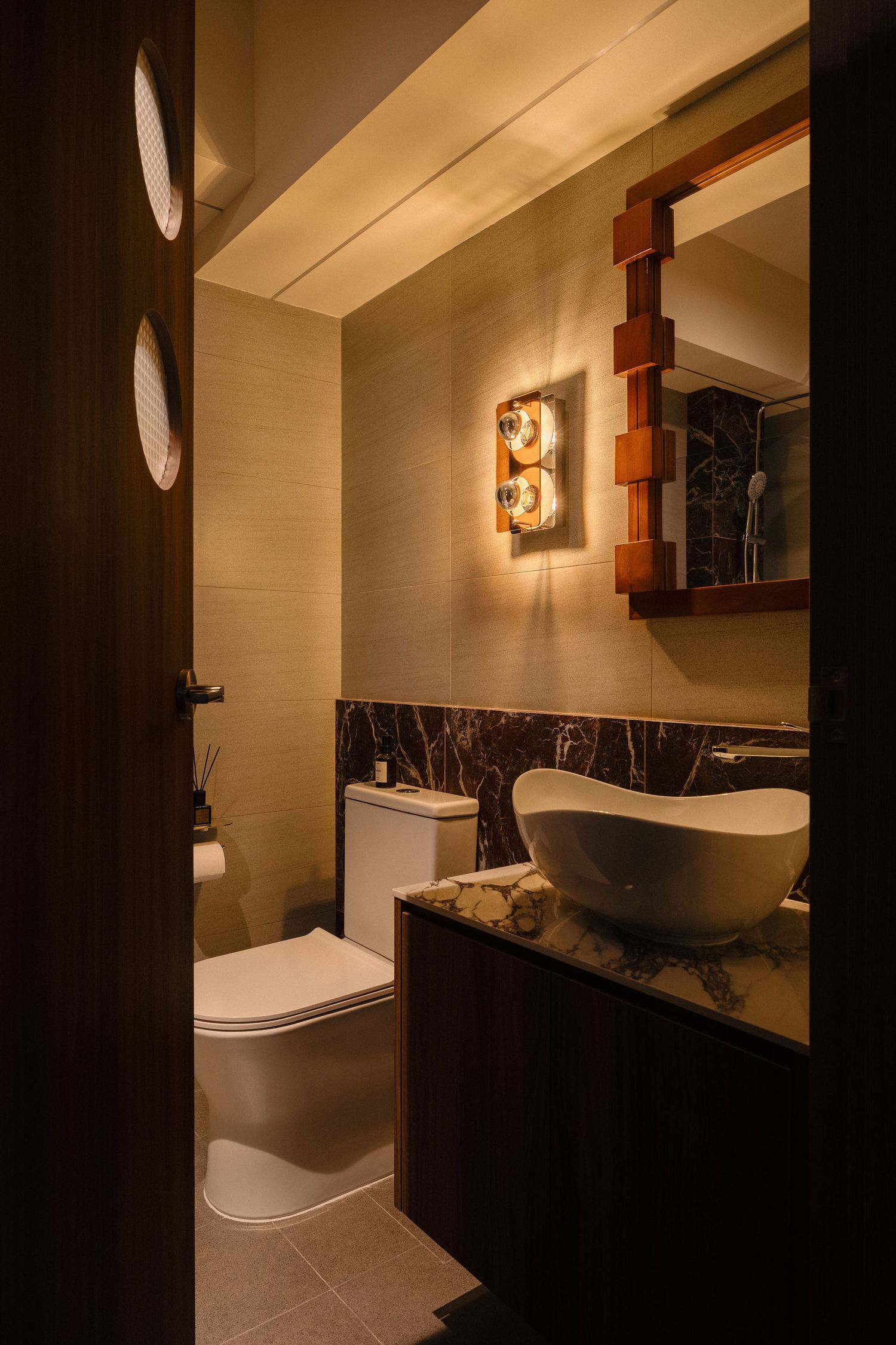 Elegant powder room with a white vessel sink, dark marble backsplash, wood-framed mirror, wall sconce and modern toilet in warm lighting.