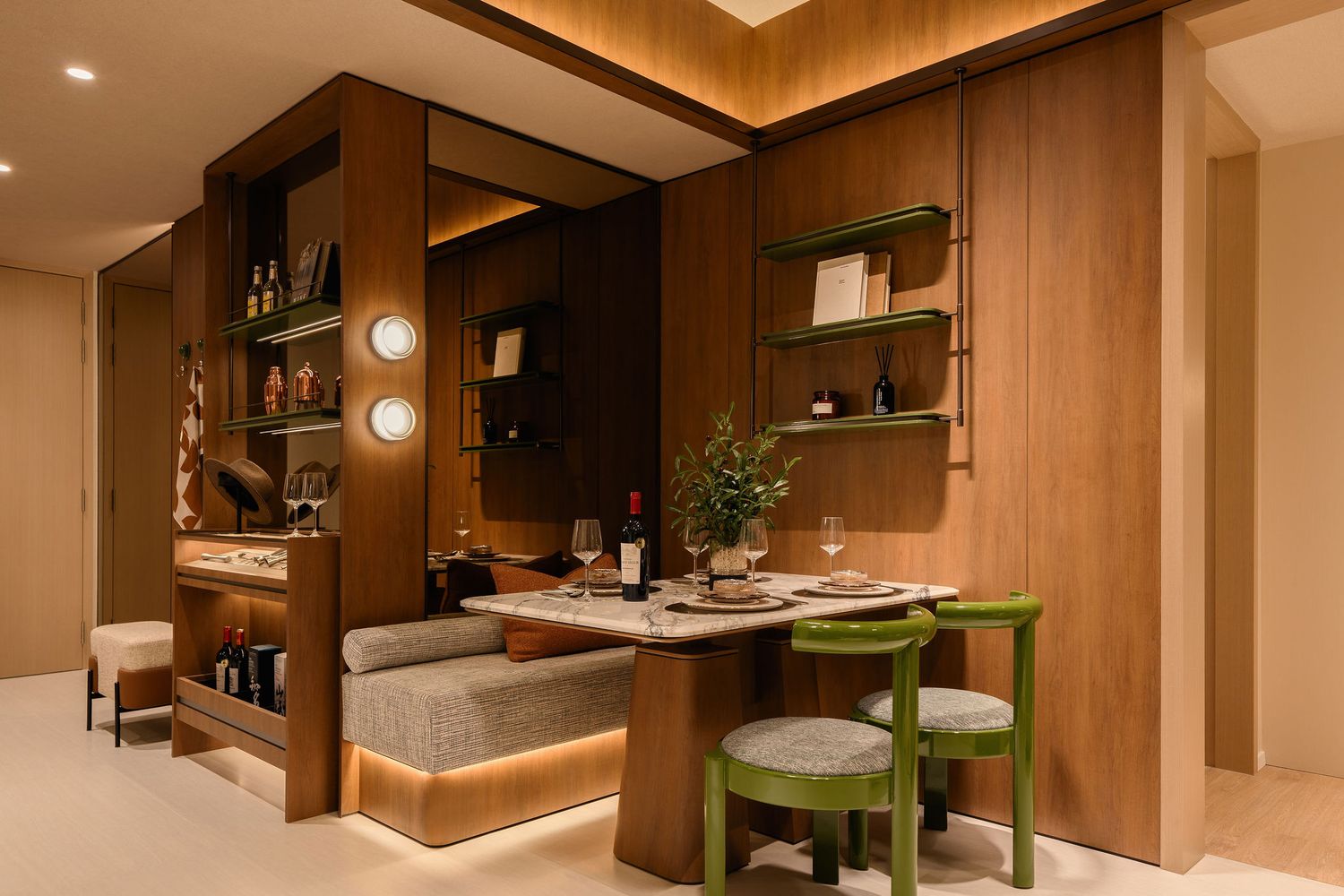 Modern dining nook with warm wood paneling, a built-in cushioned bench, green chairs, a round table with plant, and ambient lighting.