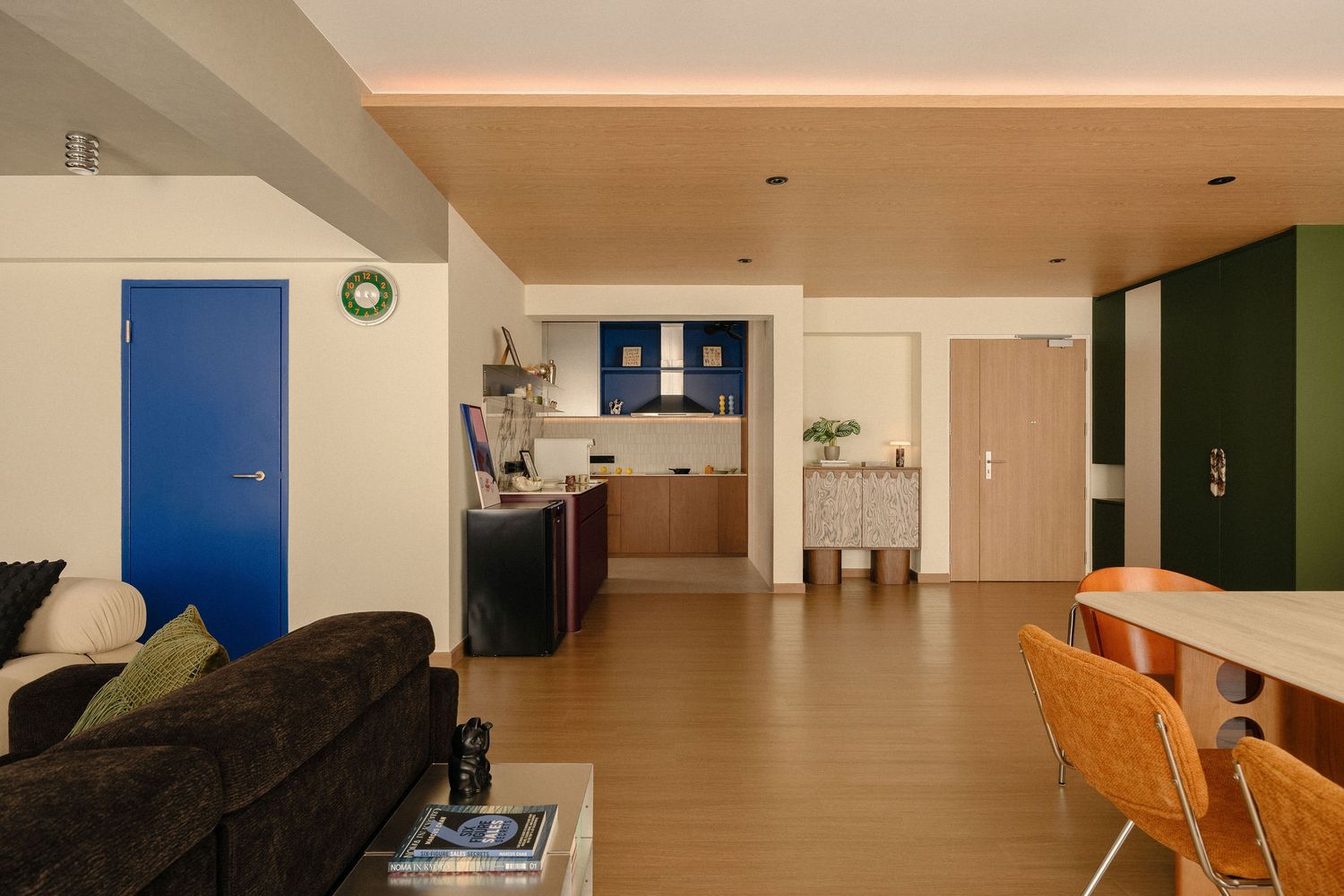 Open-plan apartment interior with a dark sofa, bright blue door, wood-paneled ceiling, green accent wall, and a kitchen in the background.