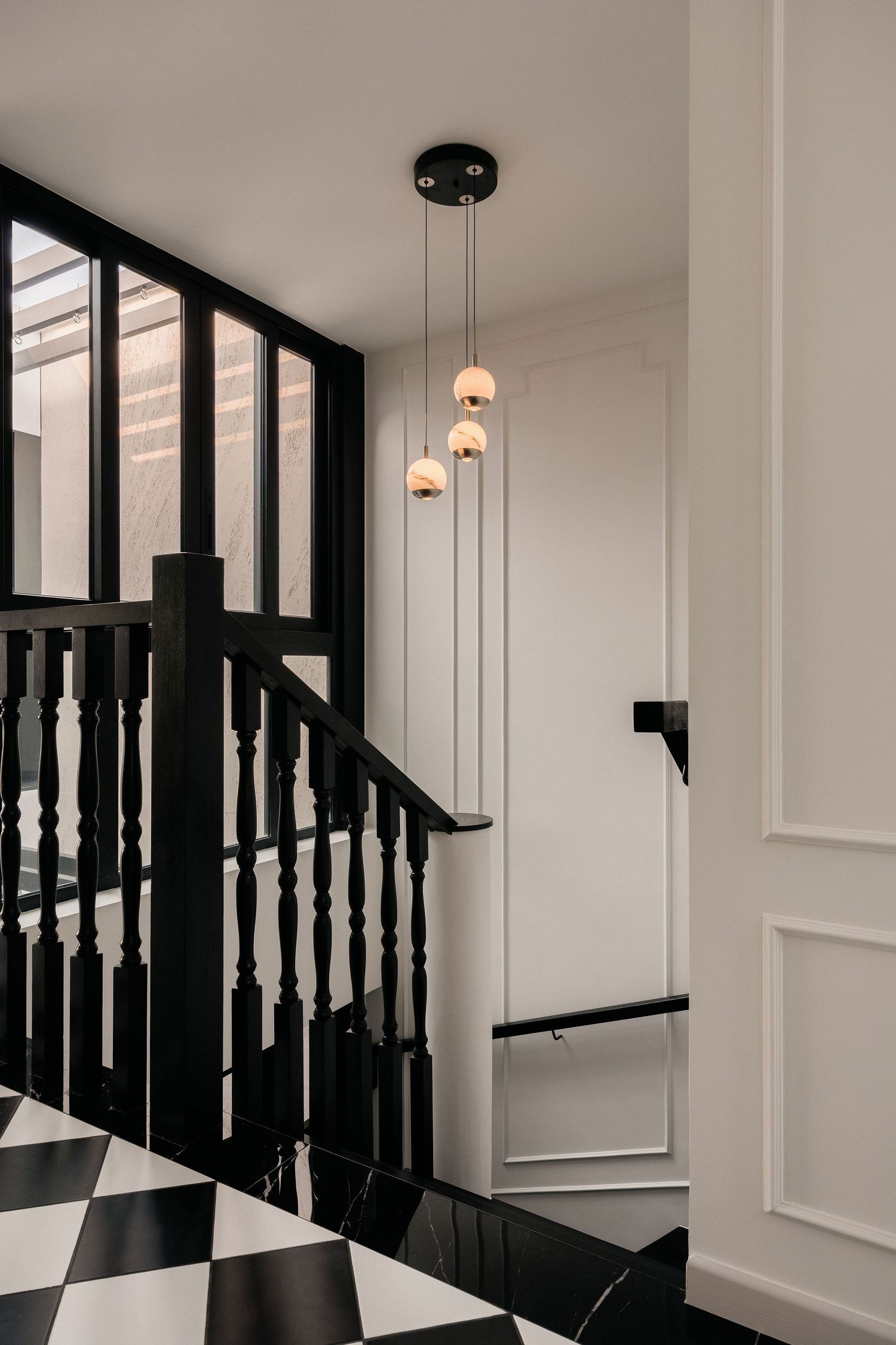 Black wooden staircase with turned balusters, white paneled walls, black and white checkered floor and a pendant light with globe bulbs.