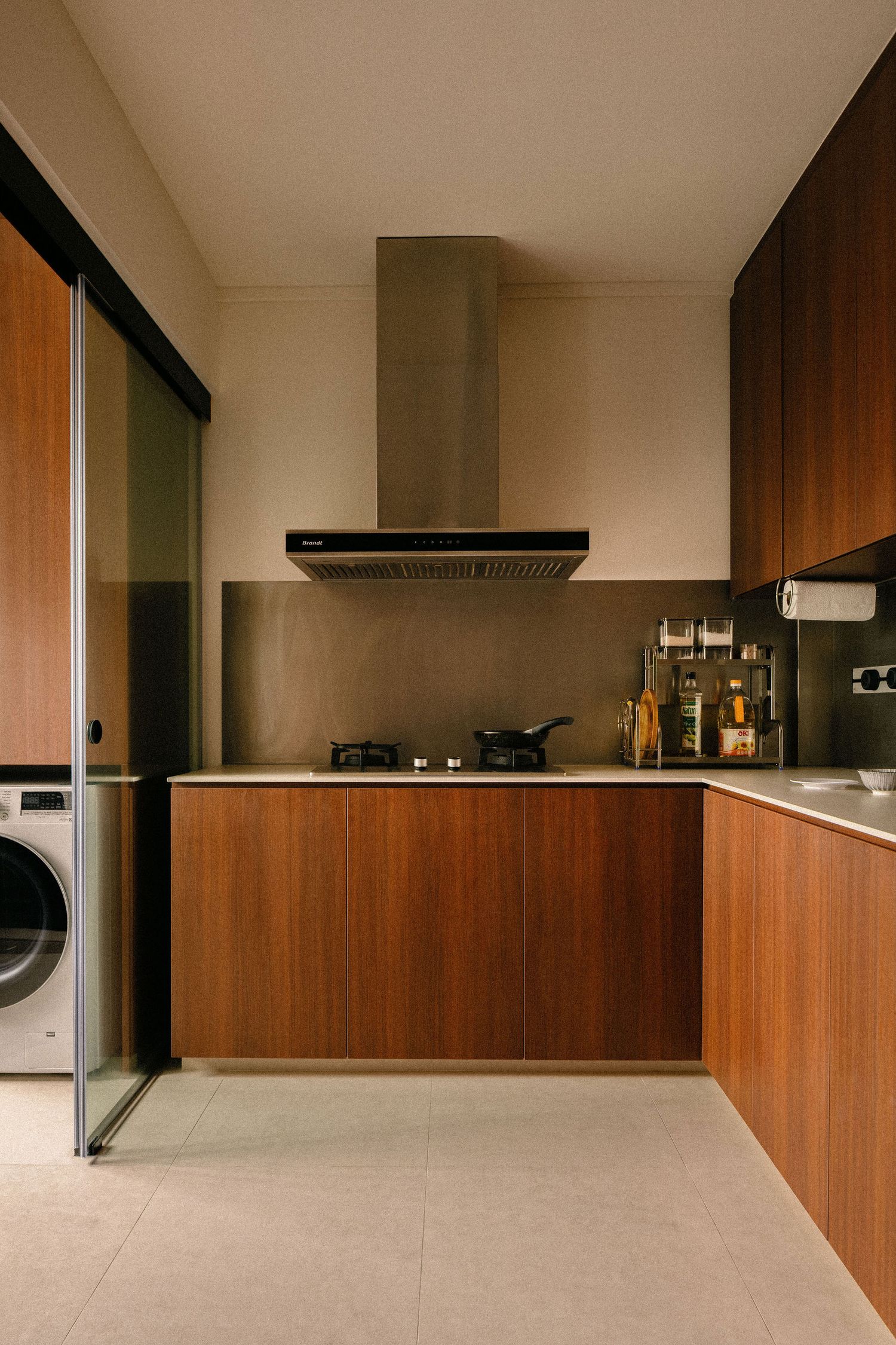 Modern L-shaped kitchen with warm walnut wood cabinets, stainless steel backsplash, gas hob, range hood and a washing machine behind a slidi