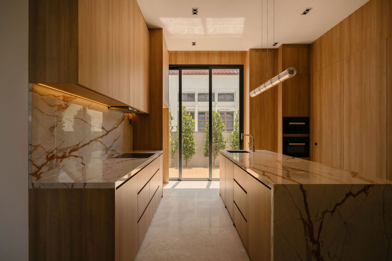 Modern galley kitchen with warm wood cabinetry, marble countertops and backsplash, and glass doors opening to a sunlit courtyard.