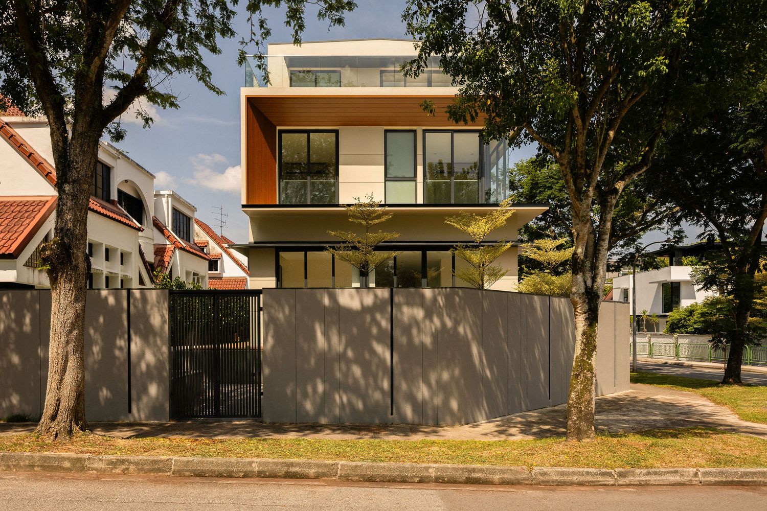 Modern three-story house with a warm wood-toned upper facade and large glass windows, set behind a gray fence and framed by mature trees.