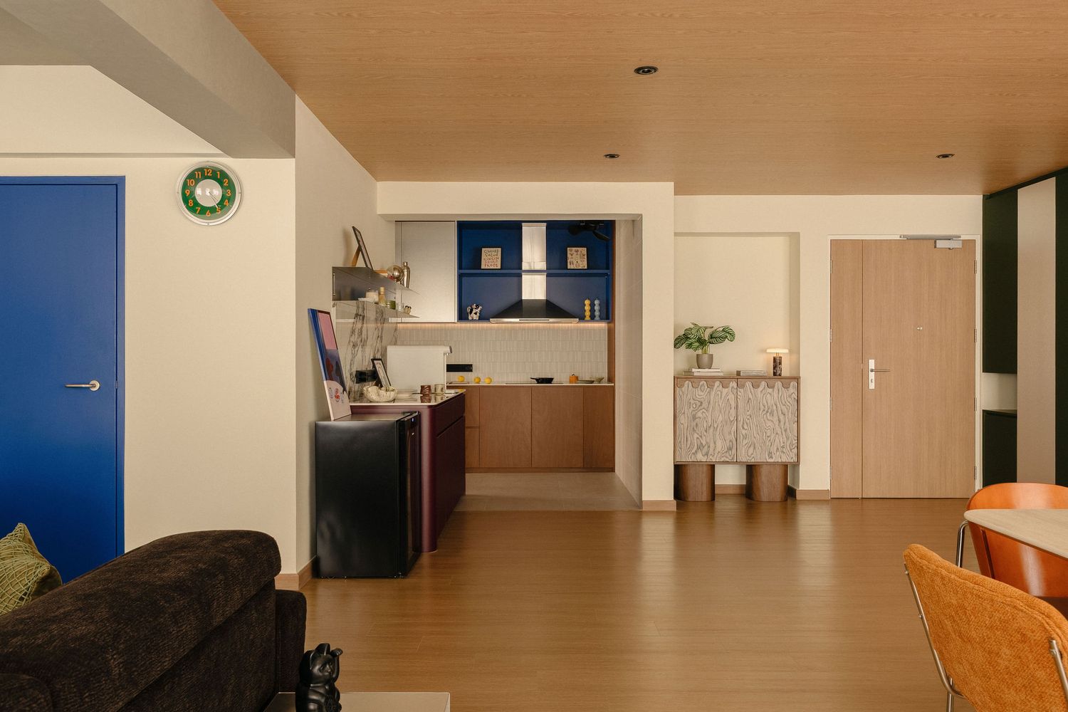 Open-plan apartment interior with warm wood ceiling and floors, a blue door, dark kitchen island, cobalt backsplash and a dining area.