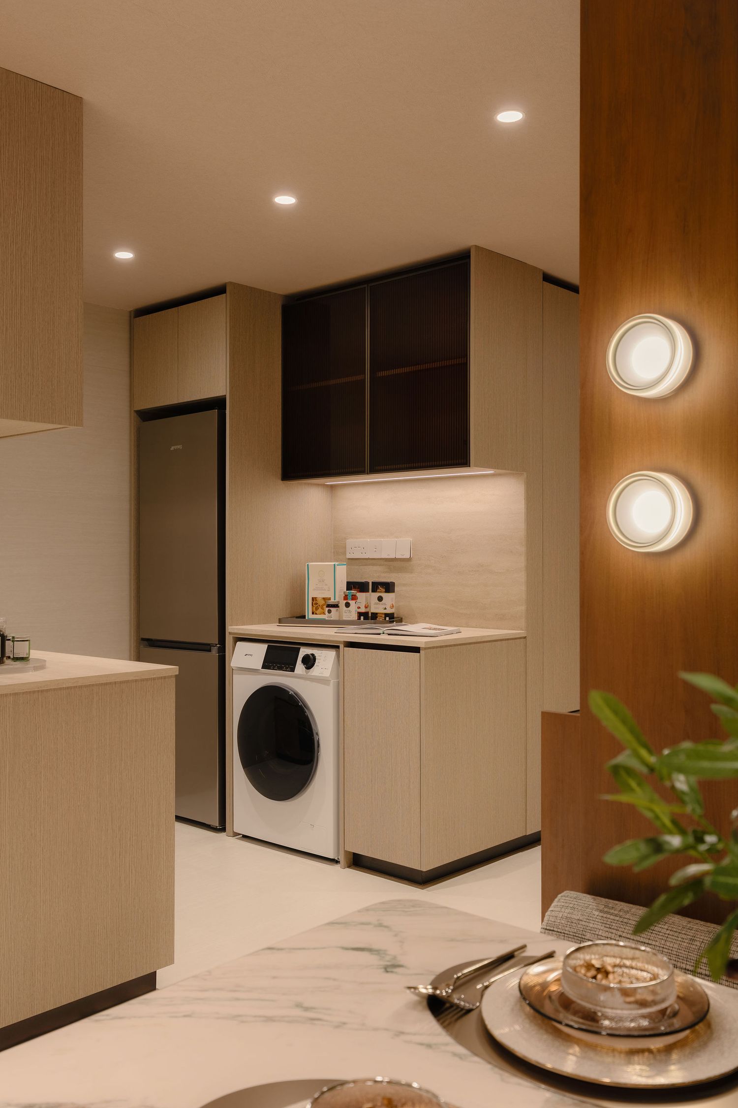 Modern compact kitchen with light wood cabinetry, built-in washing machine, tall fridge, and a marble dining table with place settings.