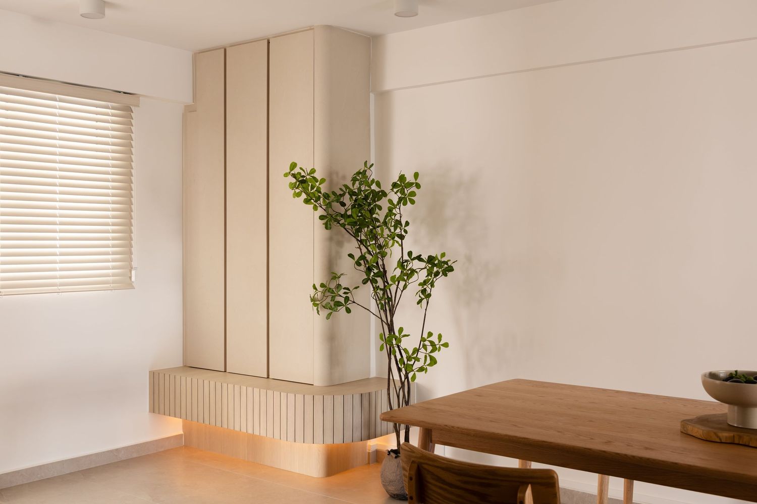 Minimalist interior design featuring a wooden dining table, beige storage unit, and potted green plant by a window.