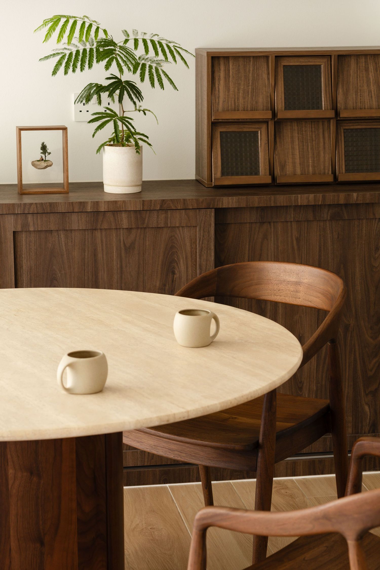Modern dining room interior with round wooden table, curved chair, and potted fern plant against walnut paneled walls.