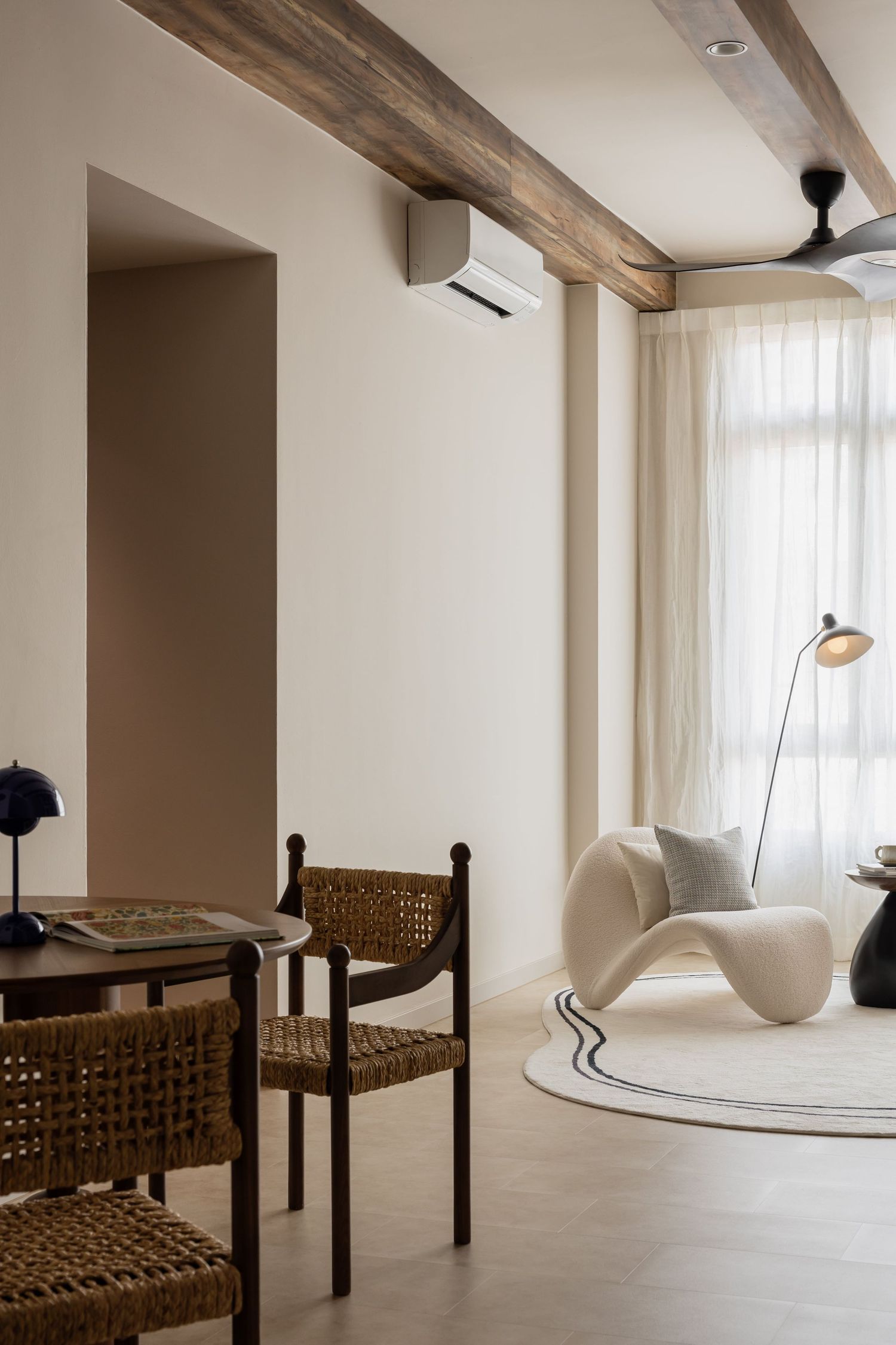 Modern living room interior with woven chairs, exposed wooden beam, white curved lounge chair and sheer curtains.