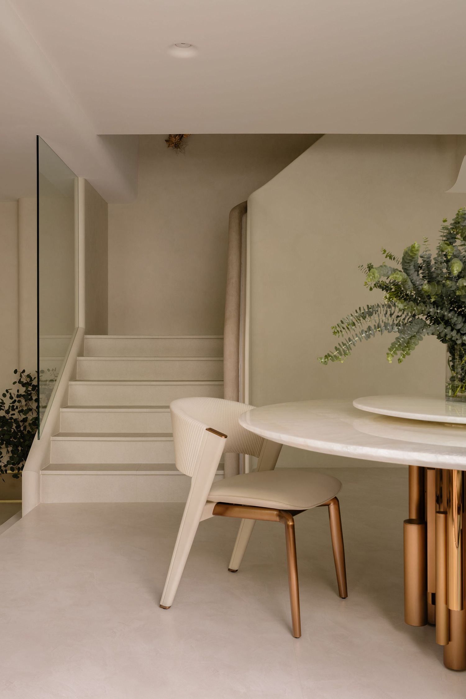 Minimalist dining area with round marble table, gold base, cream chair, and staircase in neutral-toned modern interior.