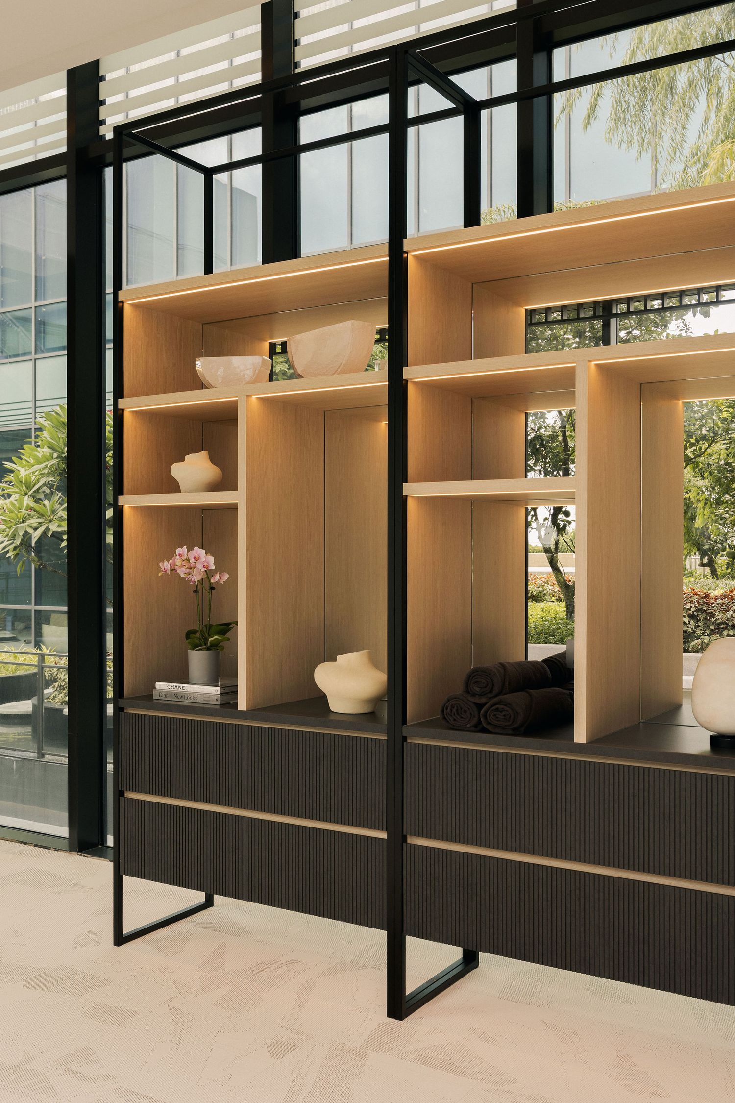 Modern open shelving unit with wood shelves, black metal frame, decorative vases, pink orchid, and folded towels near large windows.