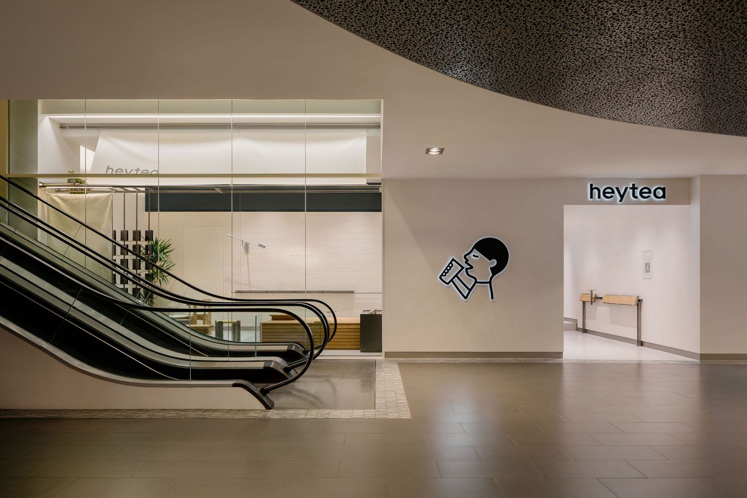 Modern mall interior with a curved ceiling, escalator, and a minimalist heytea tea shop entrance with a cartoon logo on the wall.