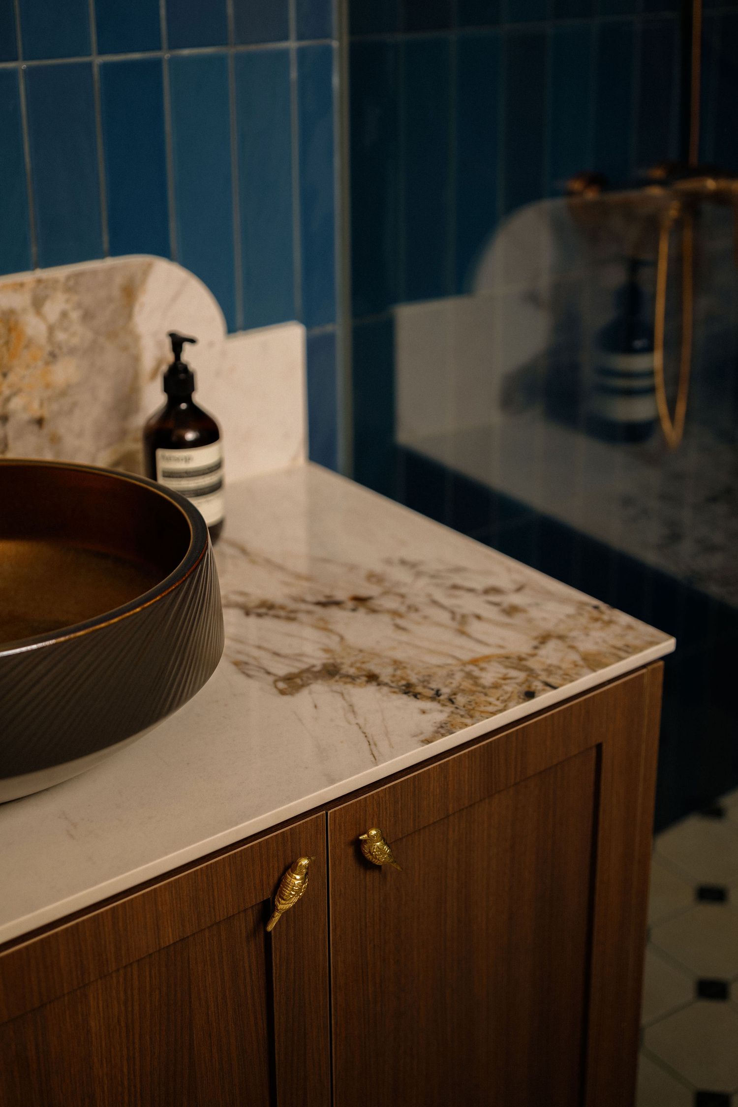 Bathroom vanity with a veined marble countertop, dark vessel sink, walnut cabinet with bird-shaped brass knobs, and blue tile walls.