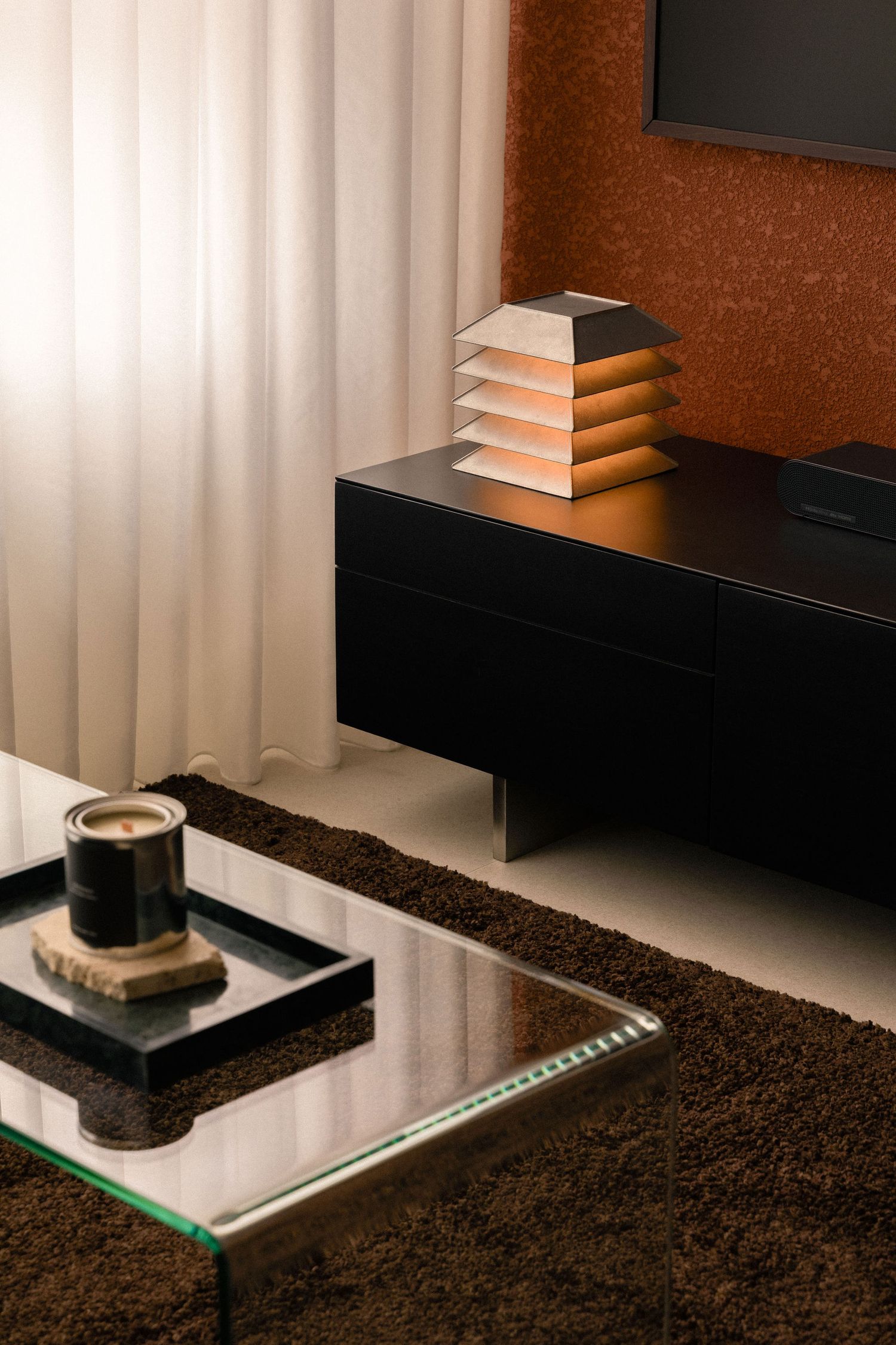 Modern living room with a glass coffee table, candle on a stone tray, black sideboard, louvered lamp, shaggy brown rug and terracotta wall.