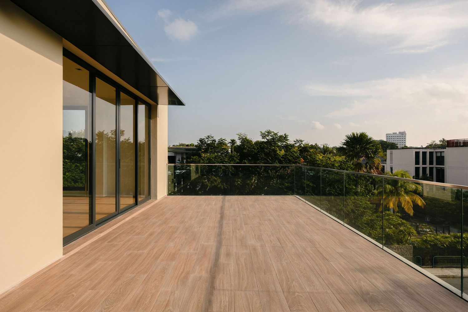 Spacious modern terrace with wood-look decking, glass railing and floor-to-ceiling sliding doors overlooking lush tropical greenery at sunse