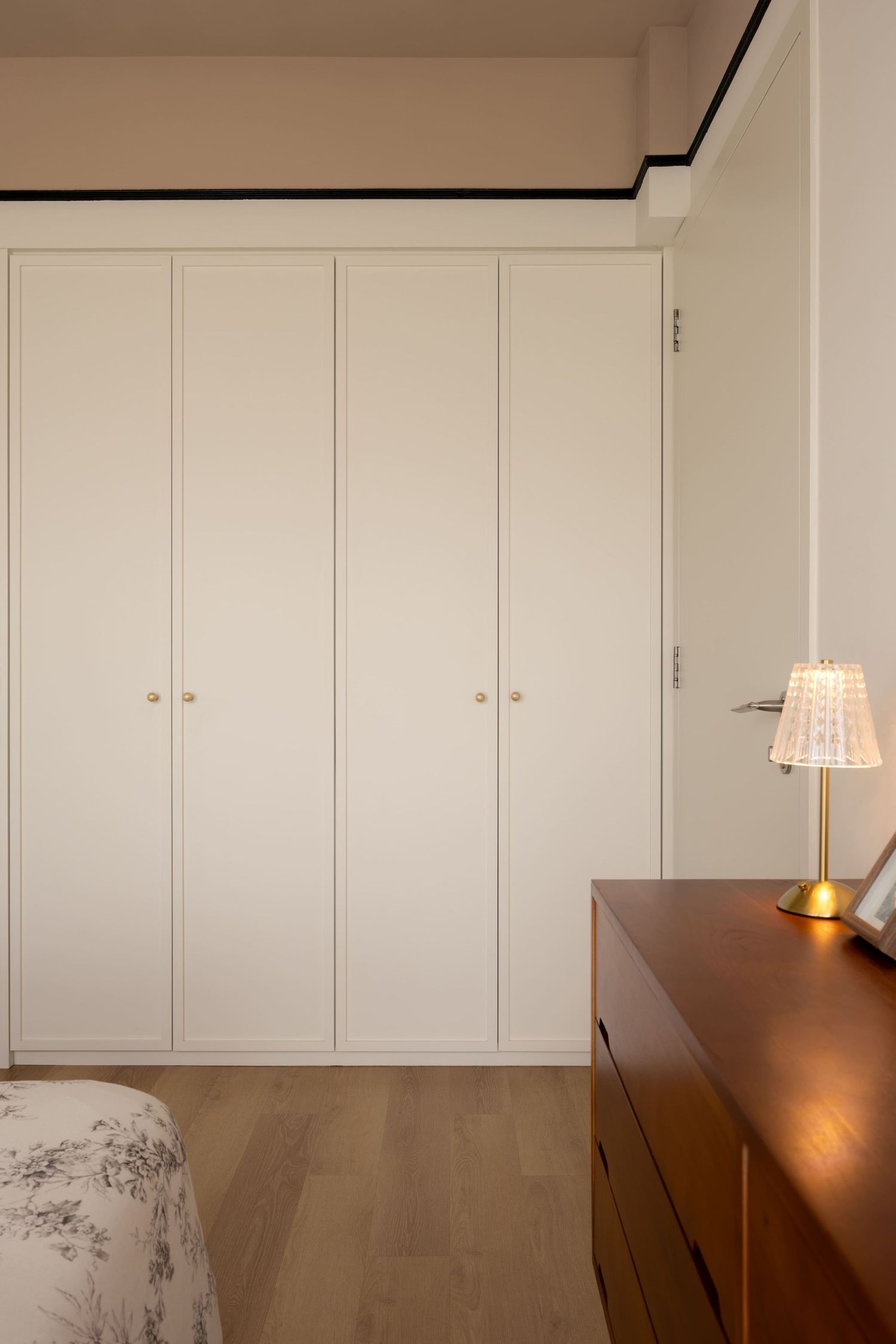 Modern bedroom with white built-in wardrobe, wooden dresser, and elegant gold table lamp on hardwood floor.
