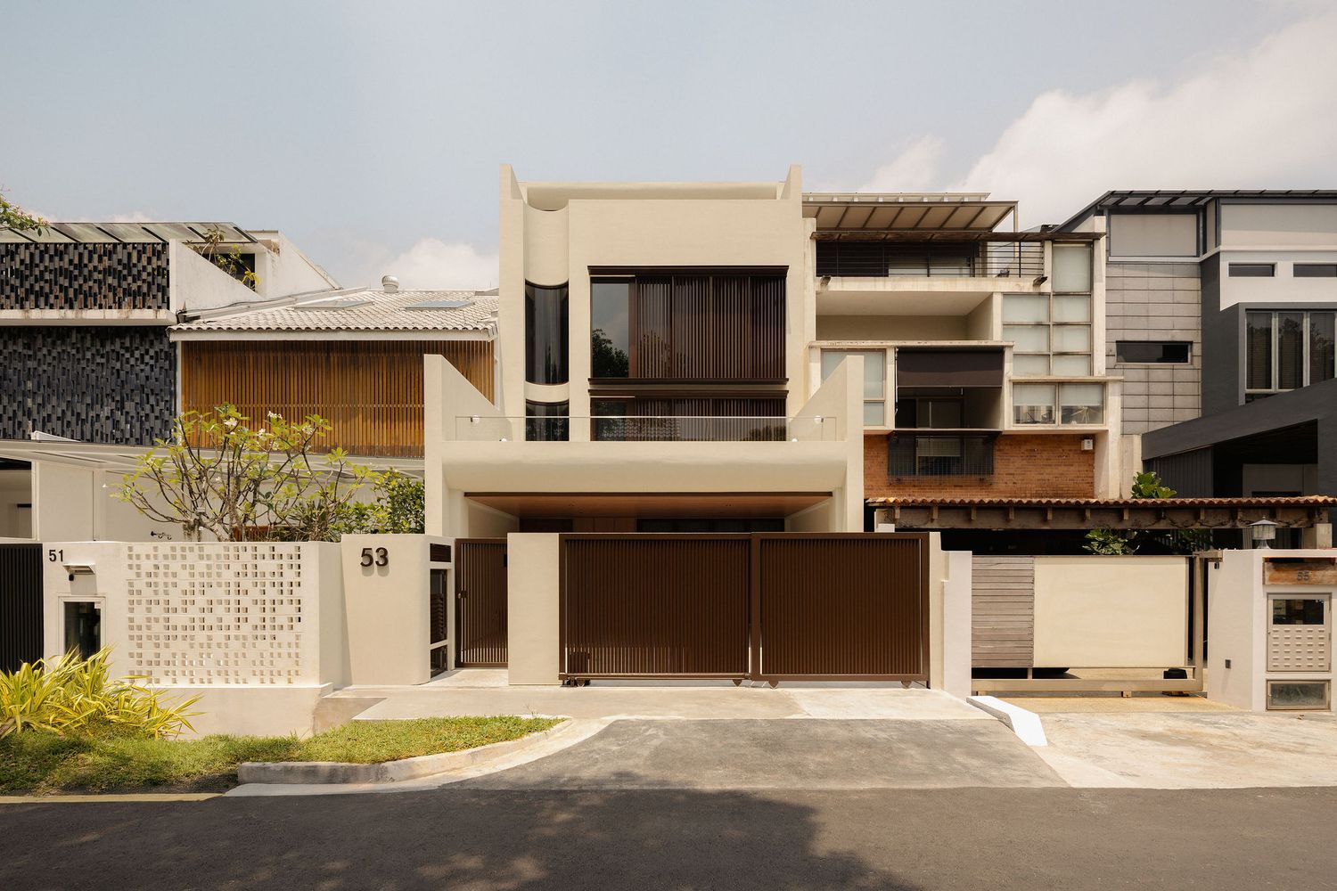 Modern two-story beige residential house with wooden garage gate, balconies, and landscaped front yard in urban neighborhood.