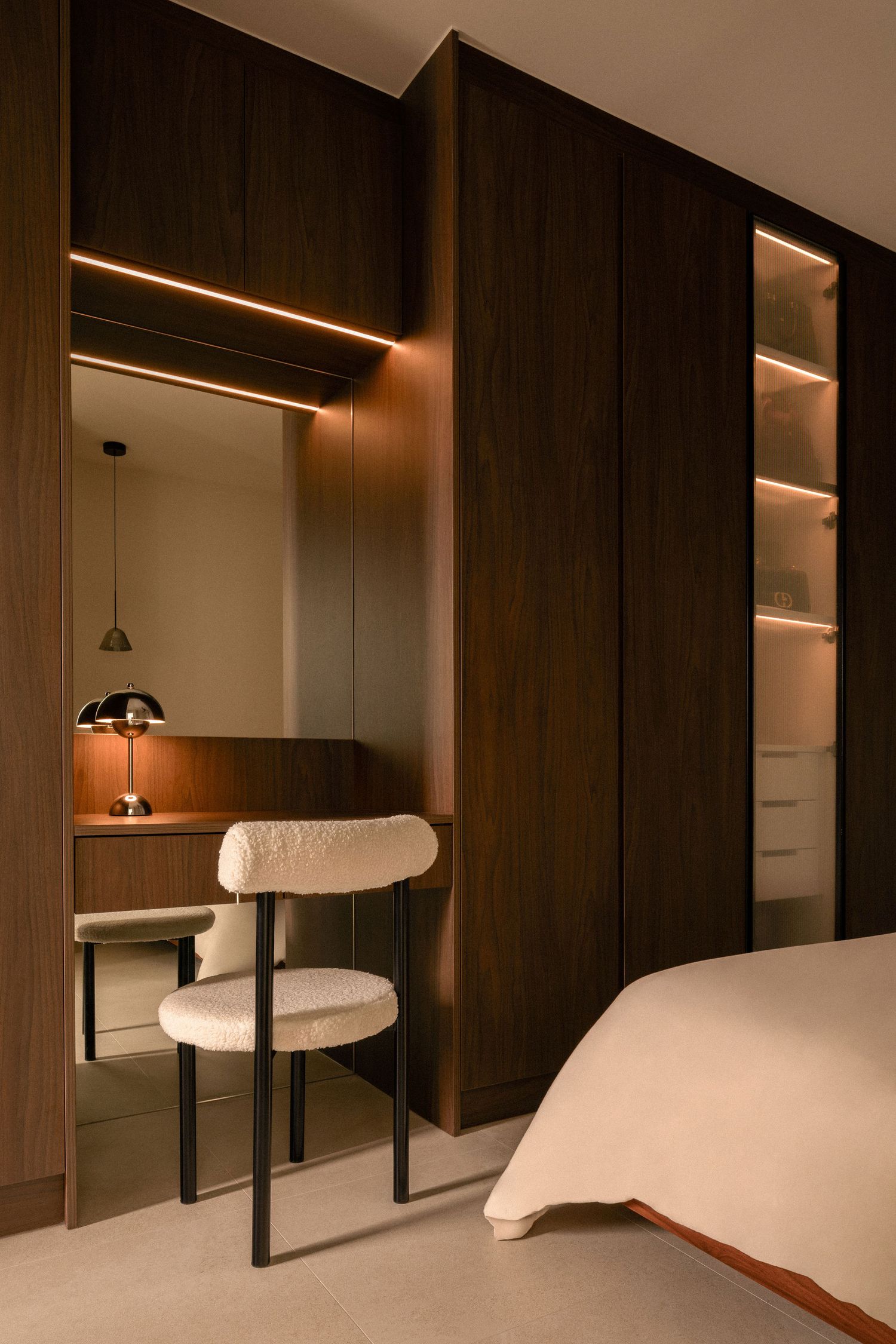 Modern bedroom with dark walnut wood cabinetry, a built-in vanity nook with warm LED lighting, a boucle chair and a cozy bed.