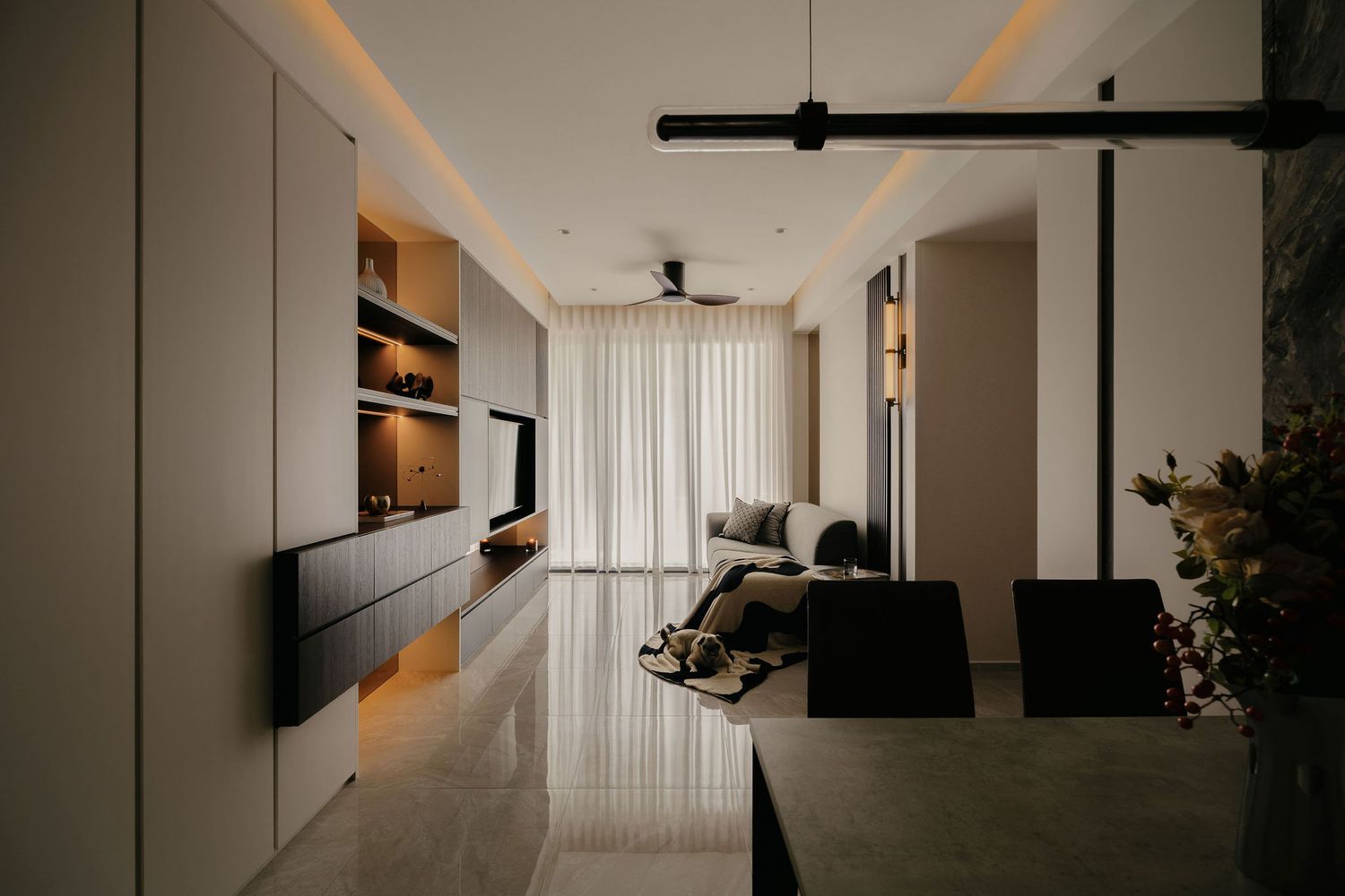Modern open-plan living room with warm cove lighting, built-in shelving, sheer curtains, a cozy sofa and a dining table in the foreground.