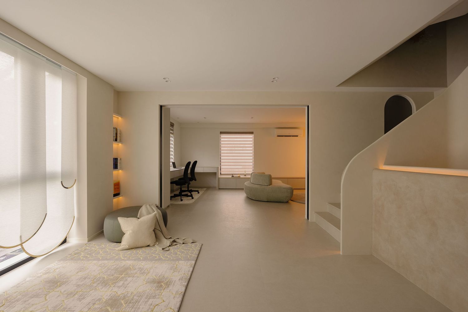 Minimalist open-plan living space with neutral tones, curved staircase, green sofa, and home office area in background.
