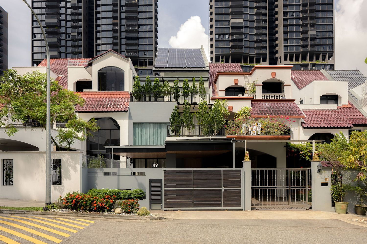 Row of semi-detached houses with red tile roofs, solar panels, and lush greenery, with modern skyscrapers in the background.