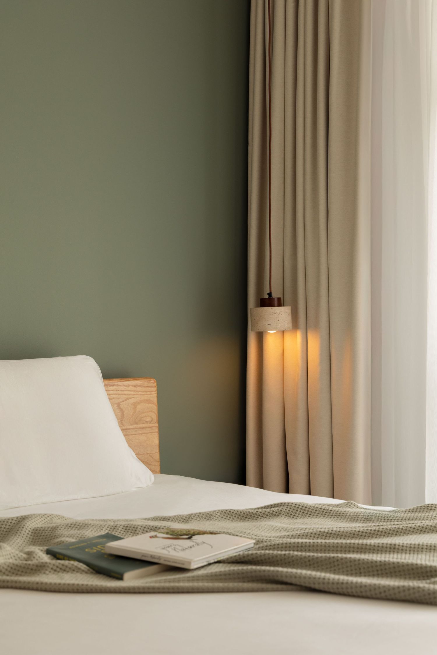 Cozy hotel bedroom with sage green walls, wooden headboard, glowing pendant lamp, neutral curtains, and a green waffle blanket.
