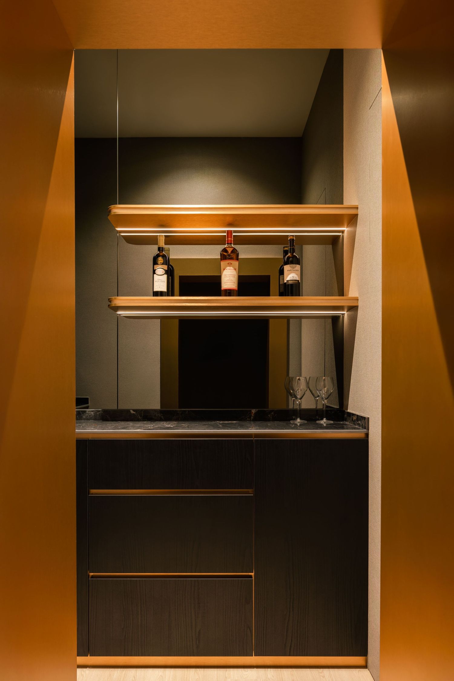 Elegant built-in mini bar with dark wood cabinet, gold shelving holding wine bottles, and two wine glasses in warm lighting.
