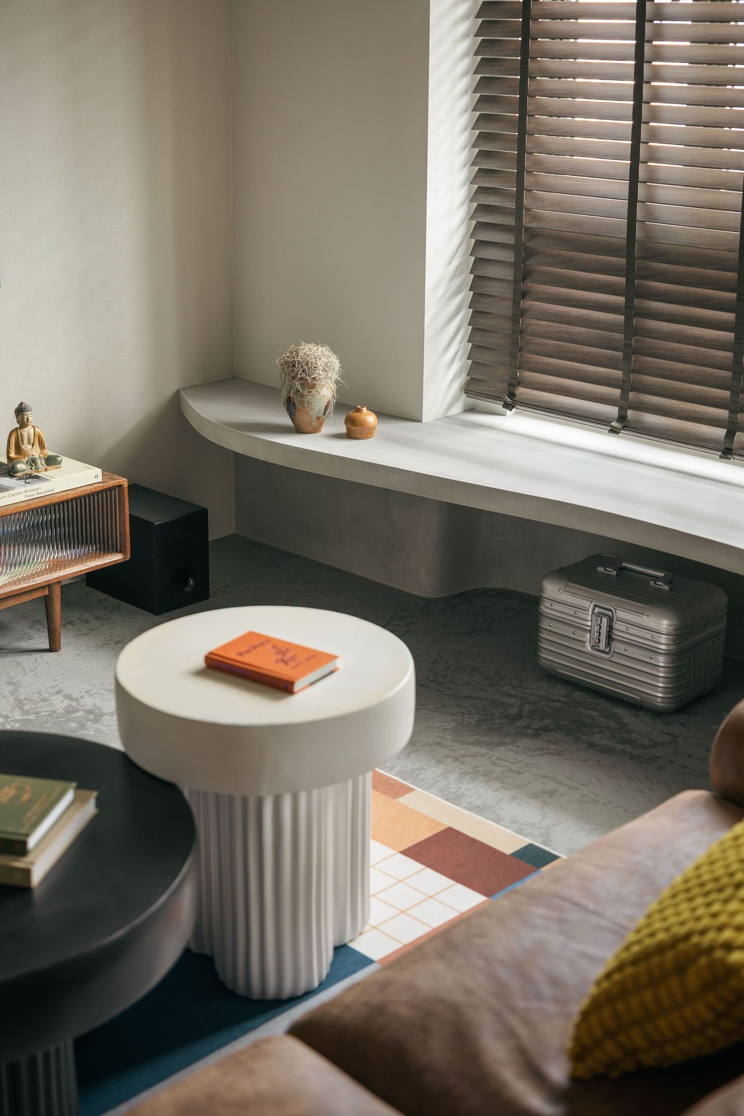 Modern living room with white fluted side table, colorful tile rug, window bench, and wooden blinds in neutral tones.