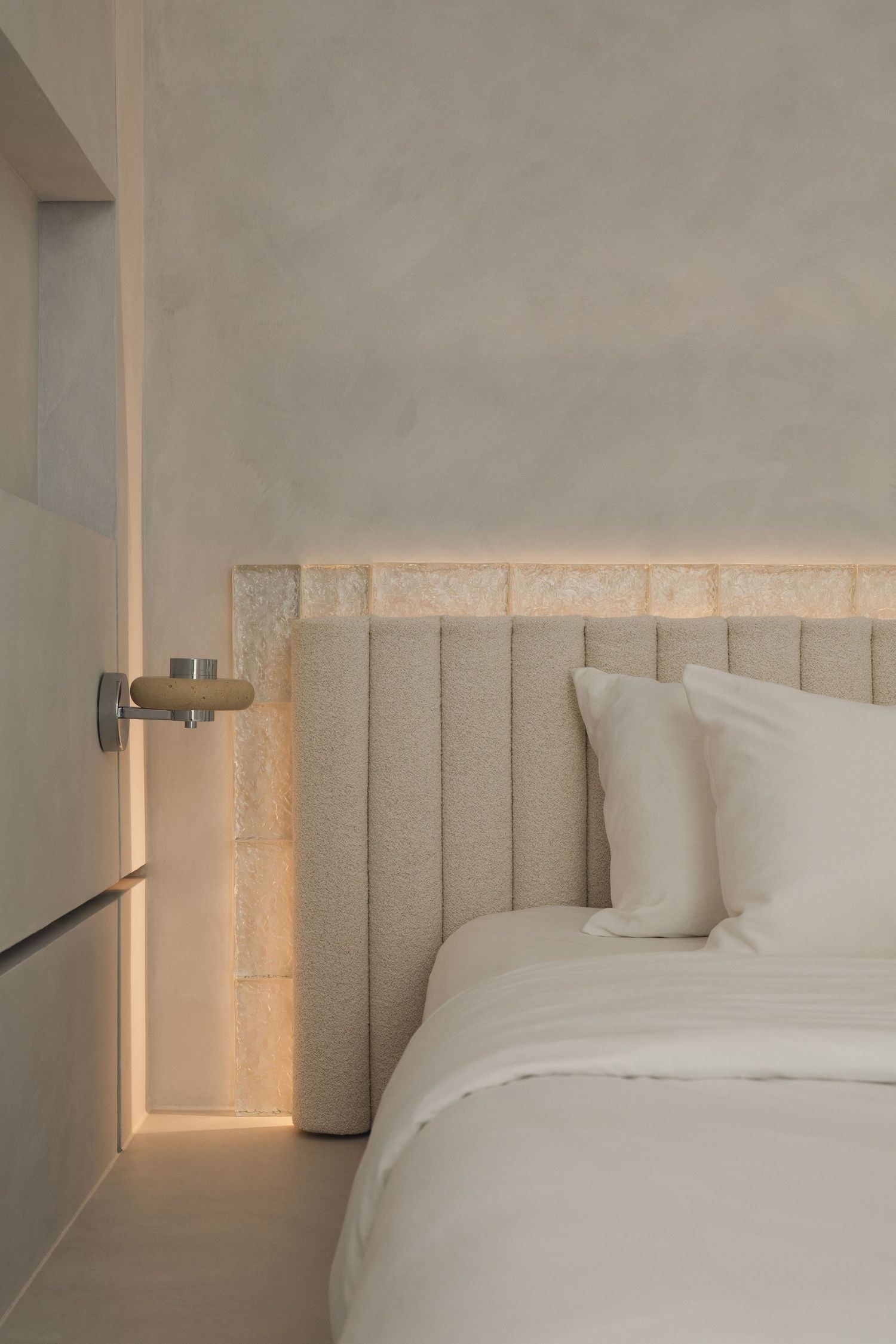 Minimalist bedroom with beige upholstered headboard, white bedding, warm wall sconce, and textured stone accent wall.