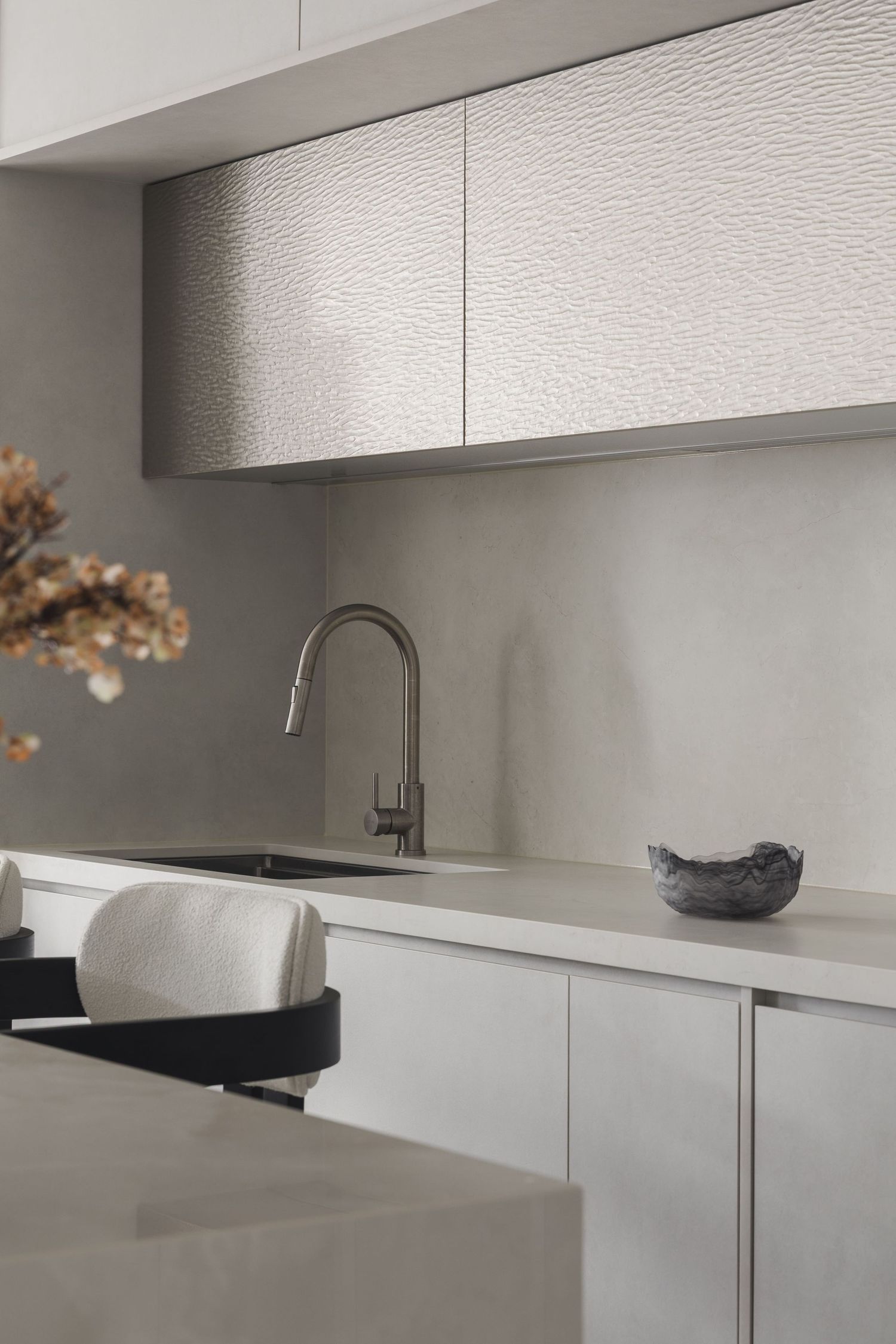 Modern minimalist kitchen with textured silver cabinets, concrete backsplash, brushed nickel faucet, and sleek grey countertops.