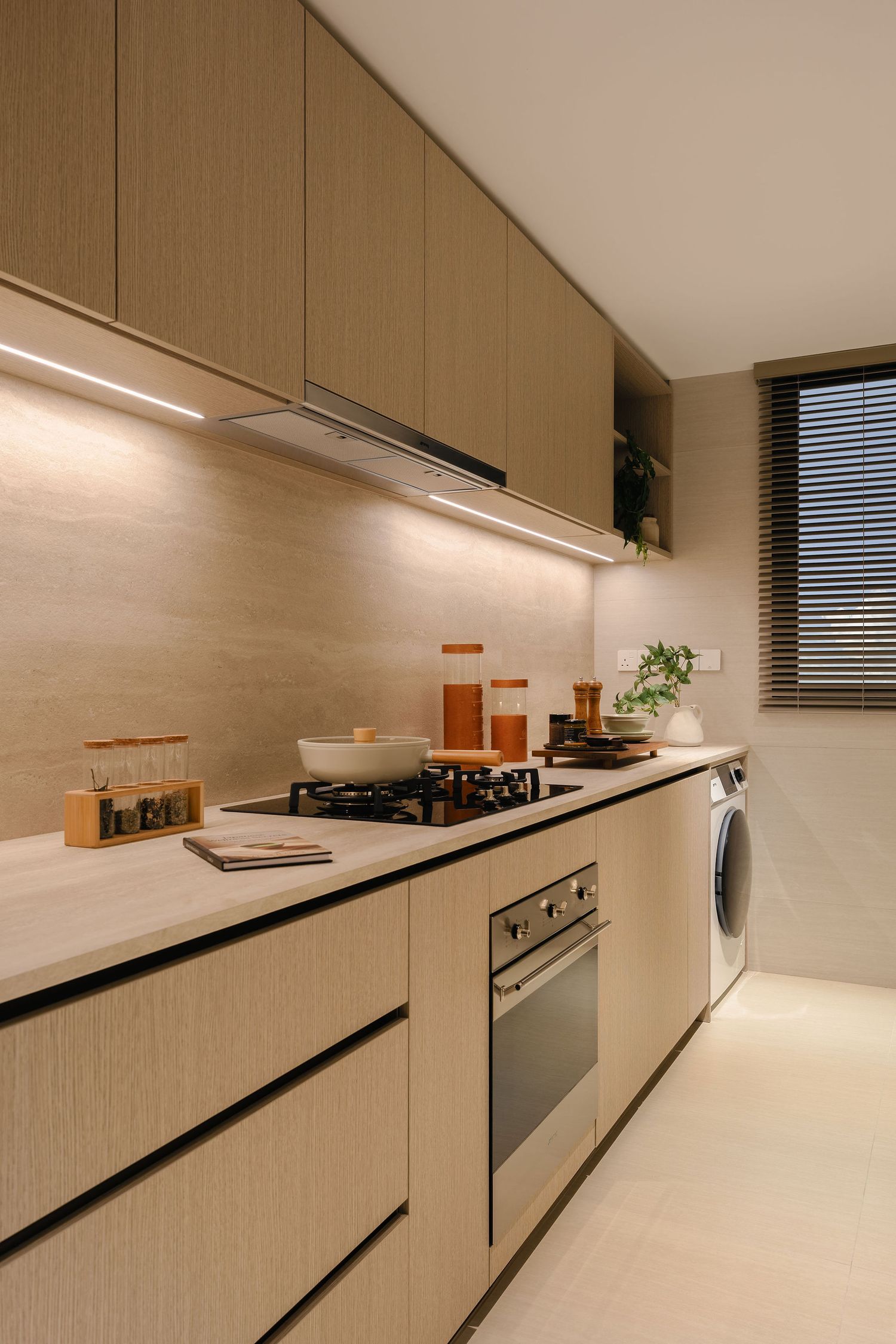 Modern galley kitchen with light wood cabinets, under-cabinet lighting, a gas hob, built-in oven, and a washing machine by a window.