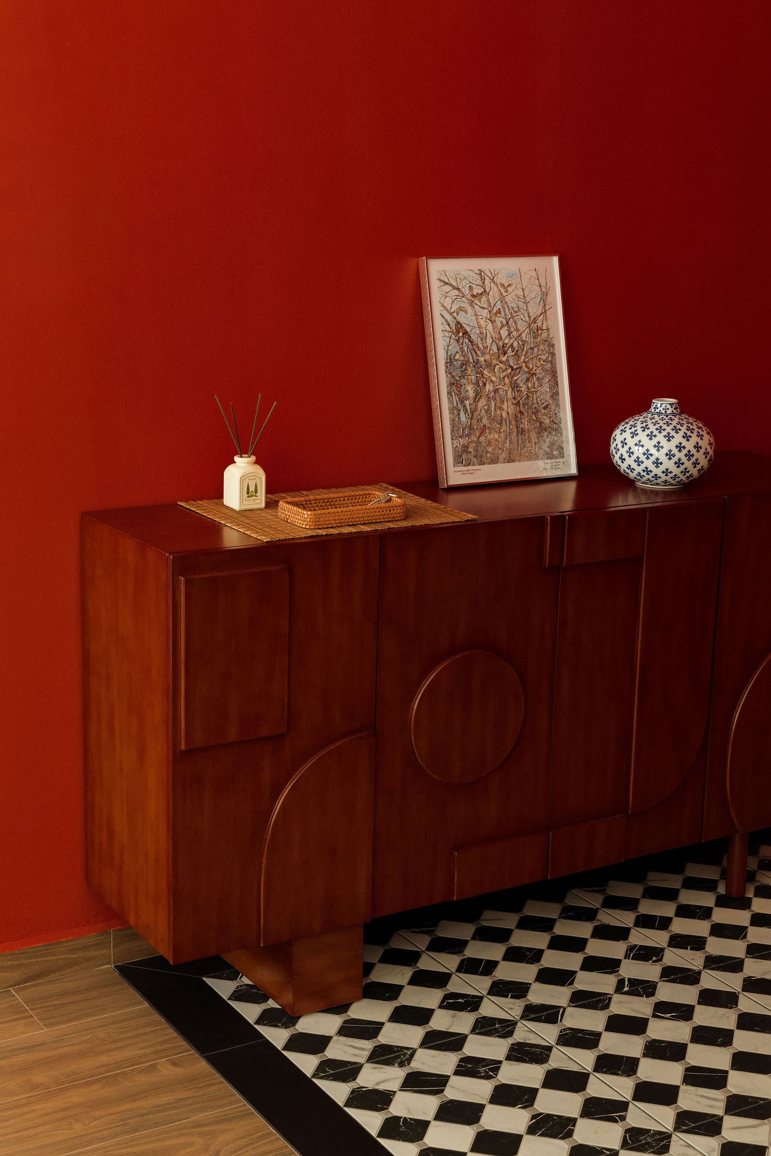 Mid-century walnut sideboard with sculpted geometric doors against a red wall, styled with framed art, a patterned vase and a diffuser.
