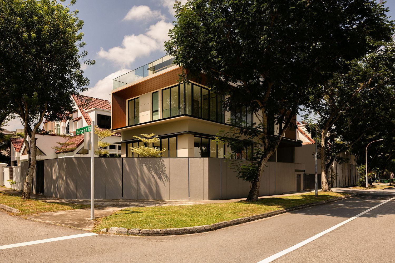 Modern two-storey corner house with glass balconies, a gray perimeter wall and mature trees along a quiet residential street.