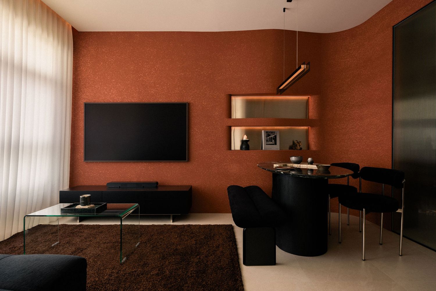 A modern living room with terracotta walls, a wall-mounted TV, black lounge chairs, a round dining table and a brown shag rug.