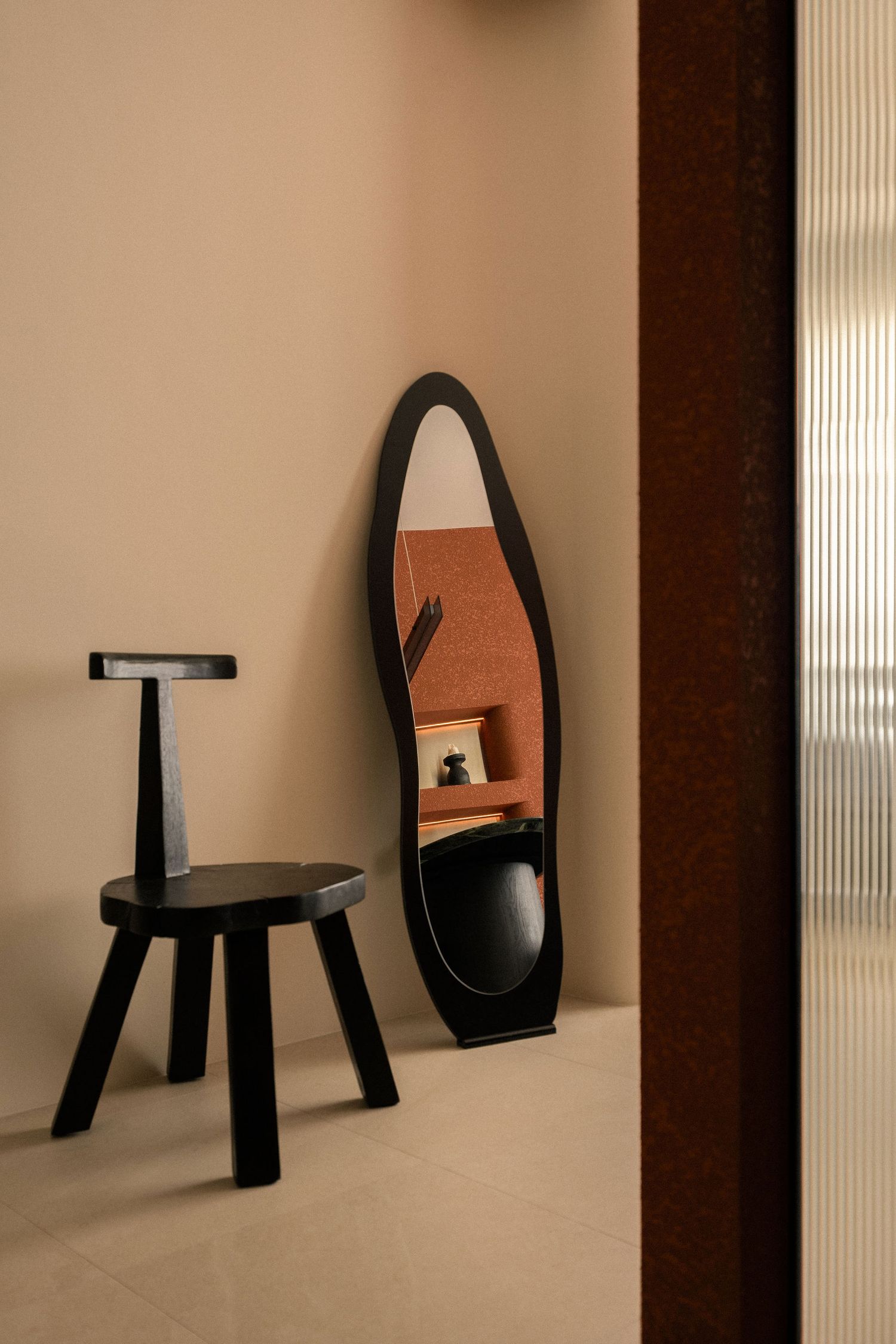 Sculptural black wooden chair with a T-shaped backrest beside an organic-shaped black-framed floor mirror in a minimalist beige interior.